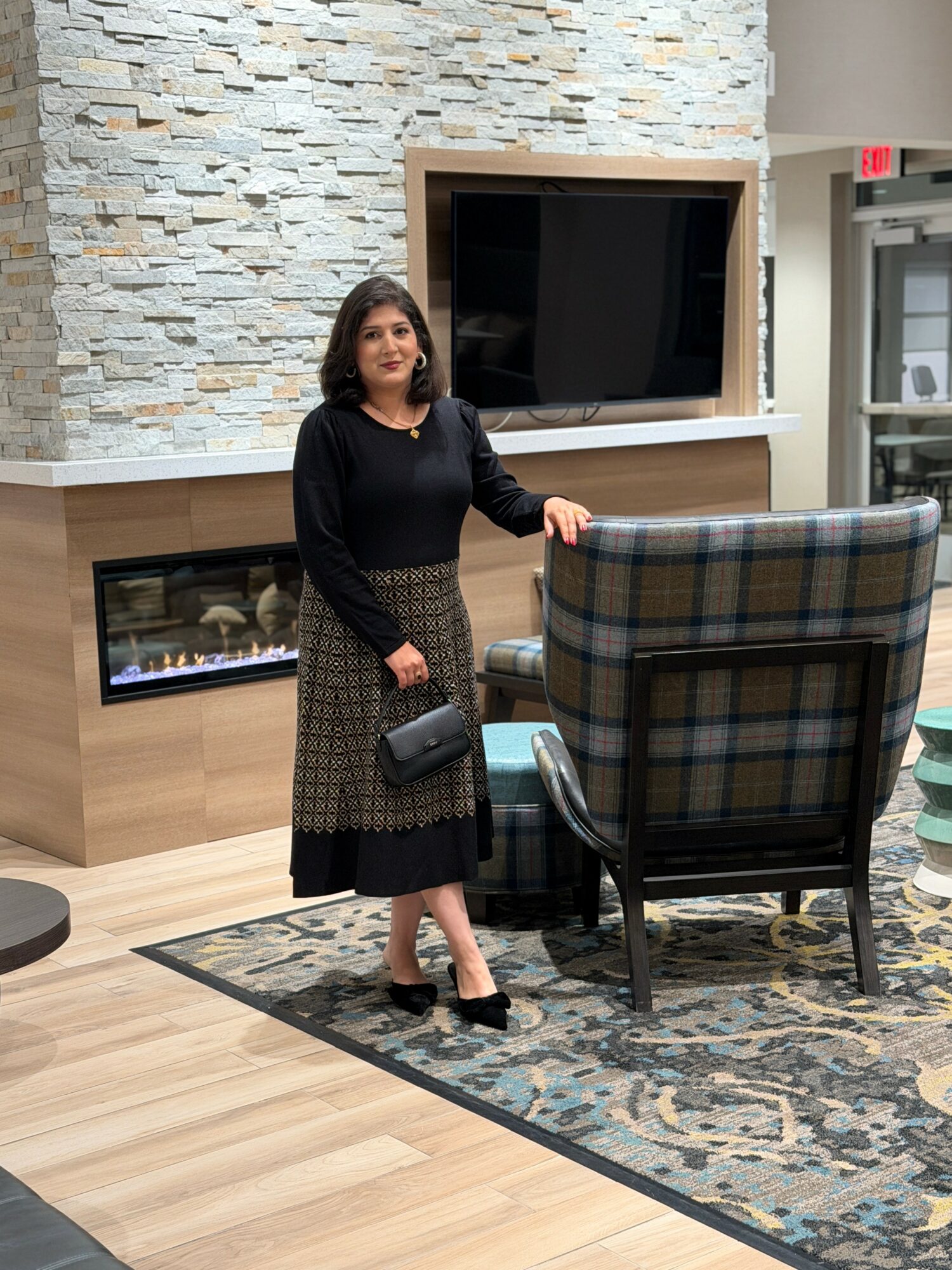 Woman standing indoors near a fireplace and large TV, holding a purse, with a plaid armchair and patterned rug nearby.
