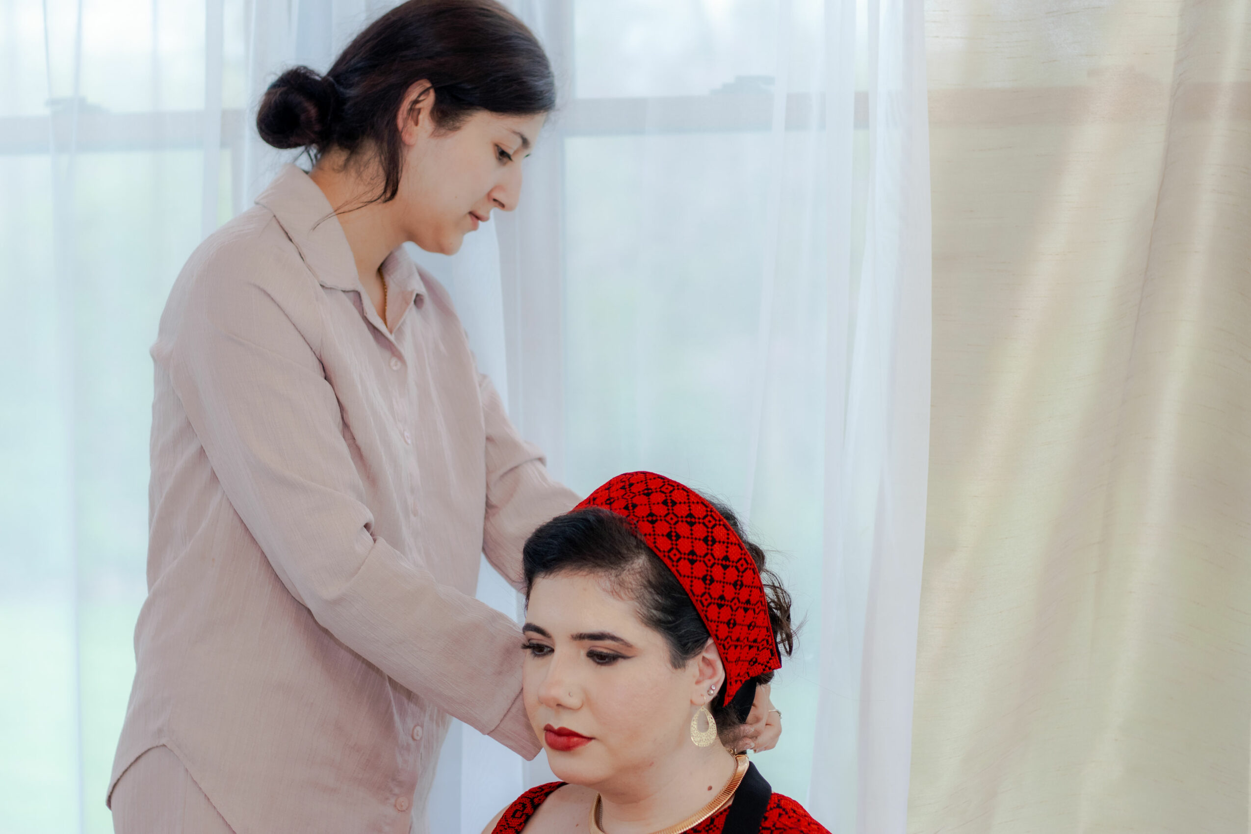 Woman with dark hair and red headscarf looking at another woman with dark hair in a beige shirt, indoors near a window.