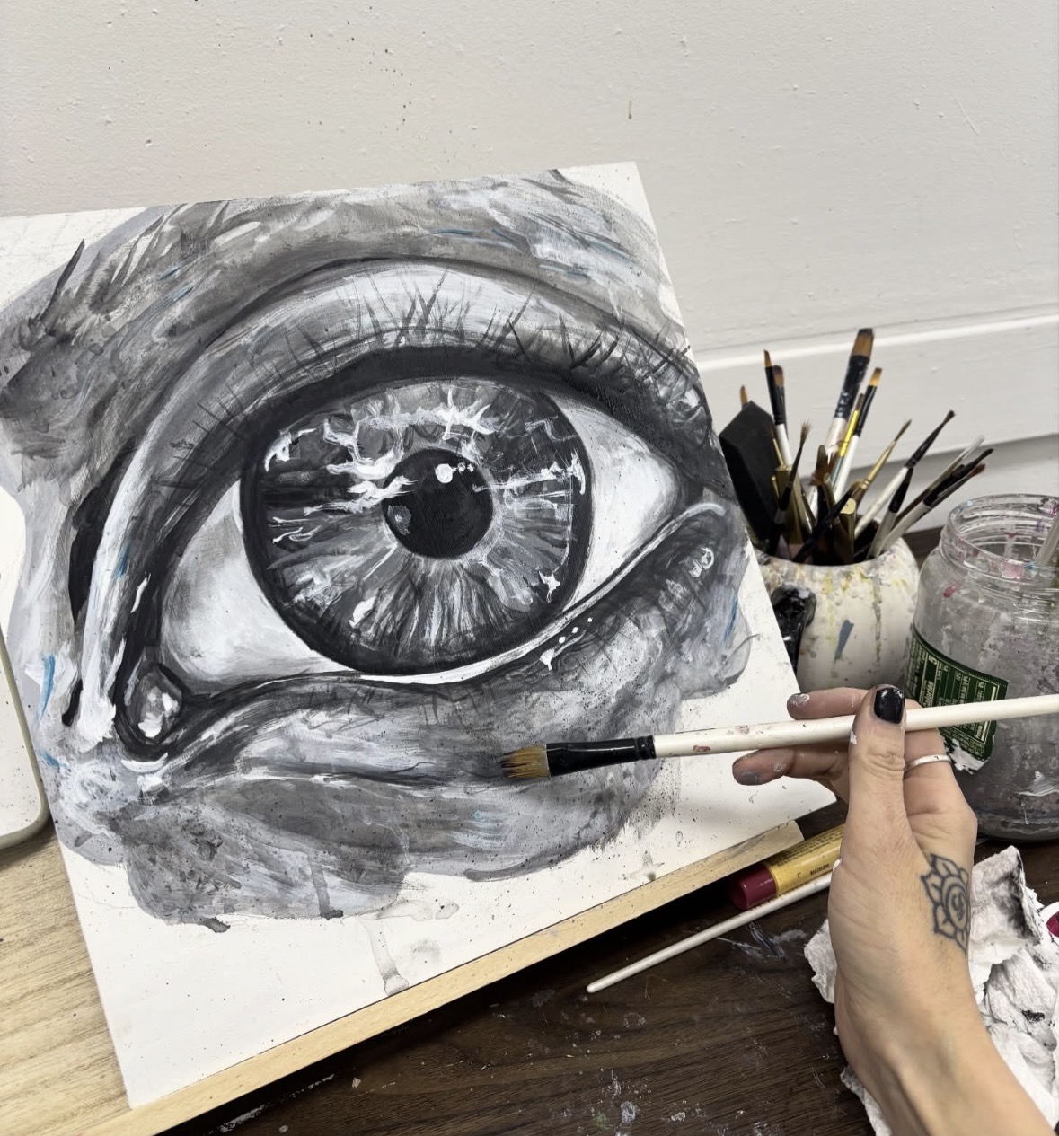 Close-up of a realistic black and white painting of a human eye on canvas, with artist's hand holding a brush nearby.