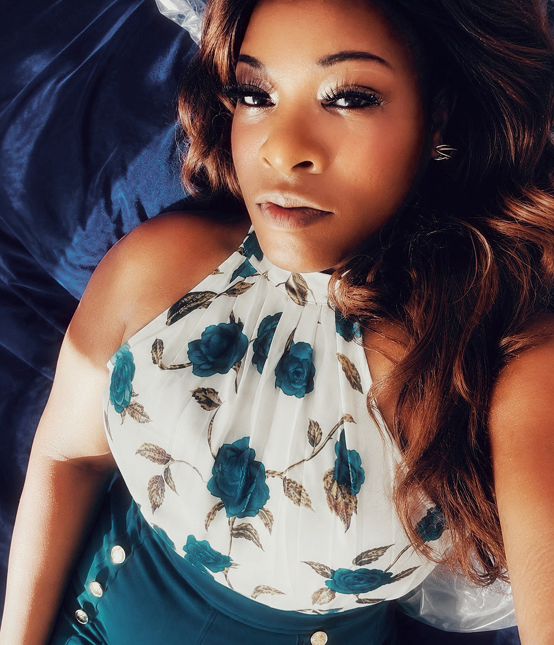 Close-up of a woman with long, wavy hair lying on a blue surface, wearing a floral top with blue roses.
