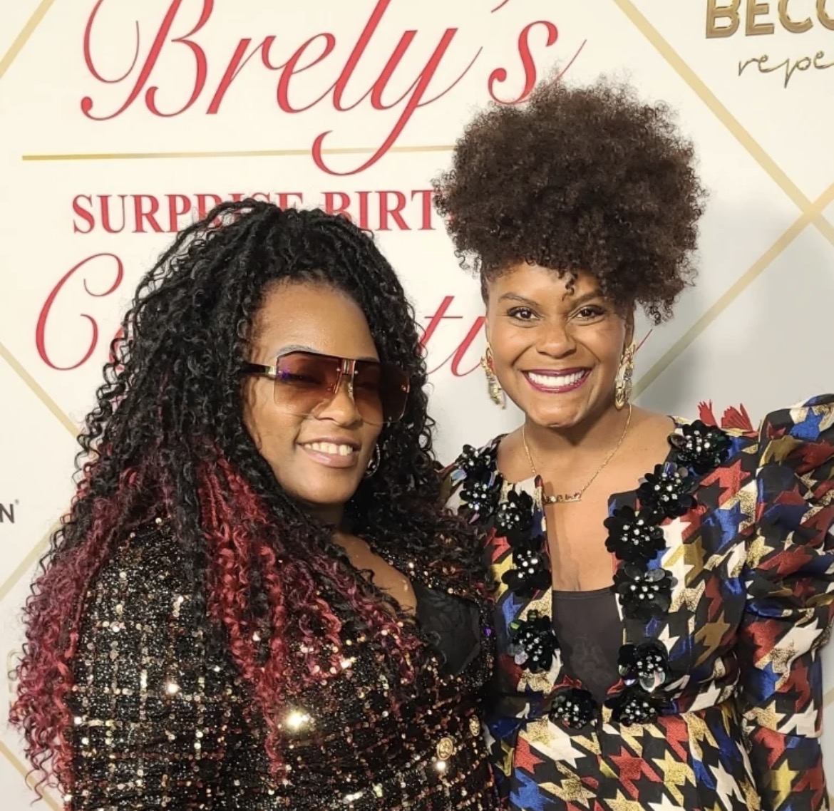 Two women smiling, one with curly hair and sunglasses, the other with an afro hairstyle, in front of a decorated backdrop.