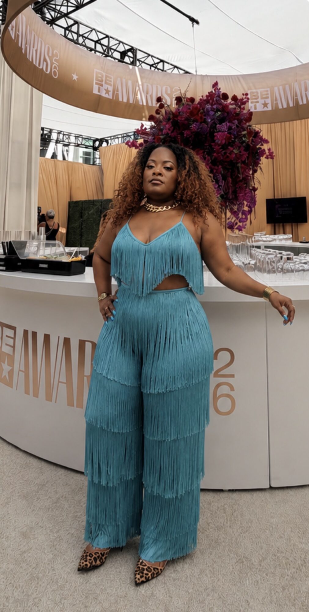 Woman in a turquoise fringed jumpsuit standing in front of a bar with a large floral arrangement behind her.