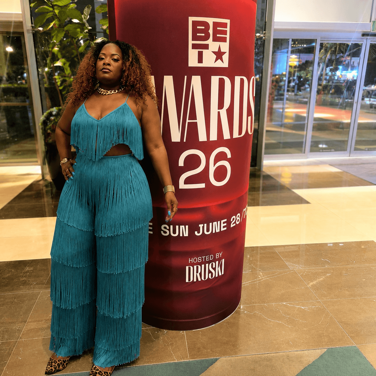 Woman with curly hair in teal fringed outfit standing next to a red BET Awards 26 sign, inside a building with glass windows.