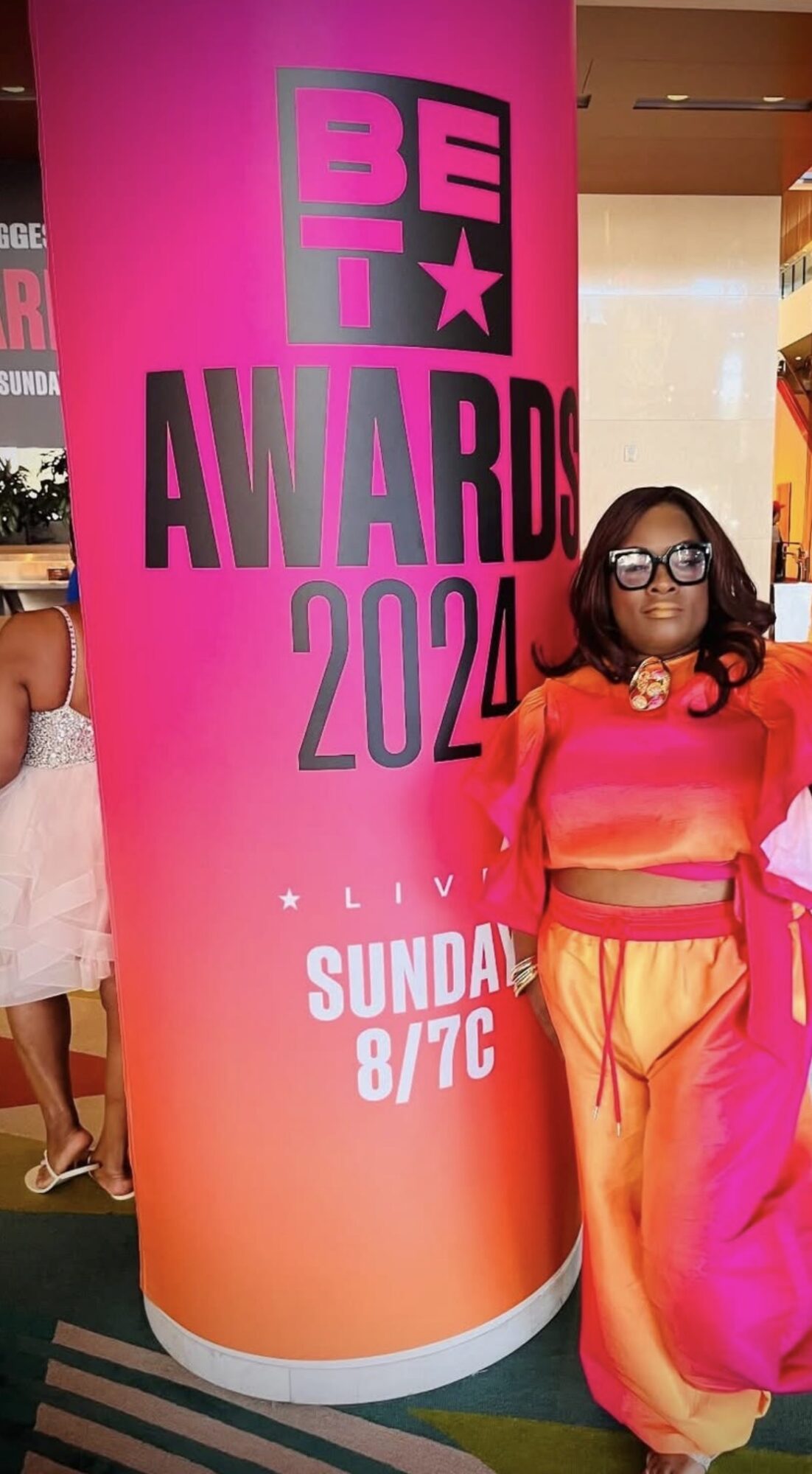 Person with glasses and colorful outfit standing next to a pink and orange BET Awards 2020 sign.