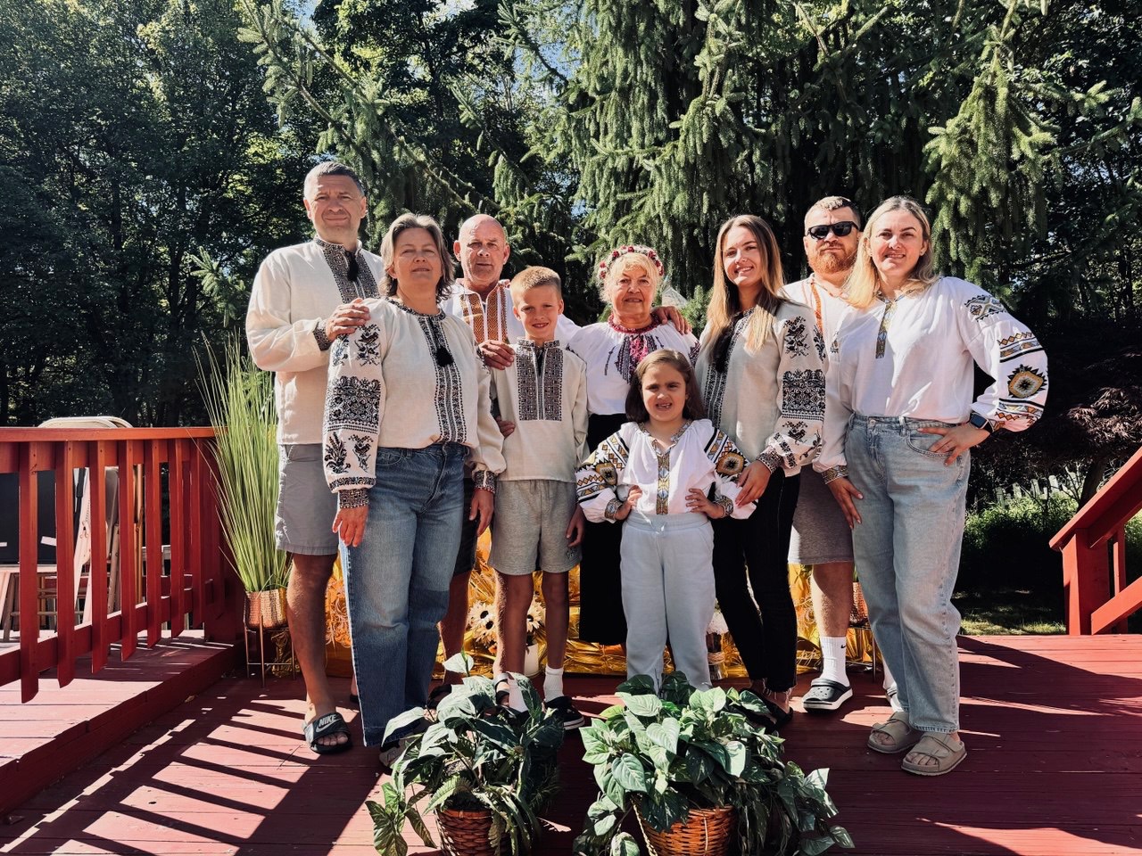 Group of nine people standing outdoors on a wooden deck with trees in the background, smiling and posing for a photo.