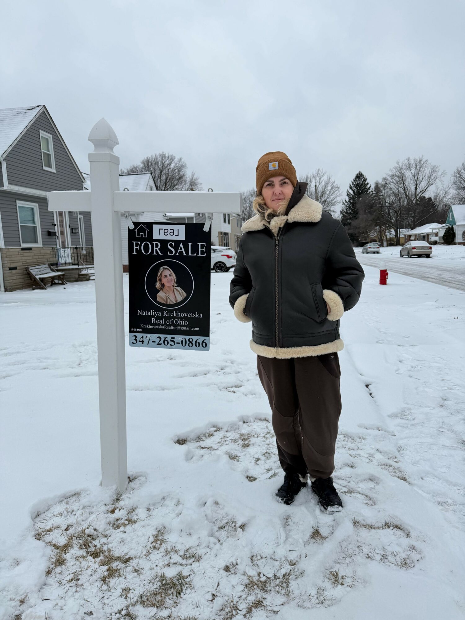 Person standing outdoors in snow, wearing a black jacket with beige collar, brown pants, and black shoes, holding a phone.