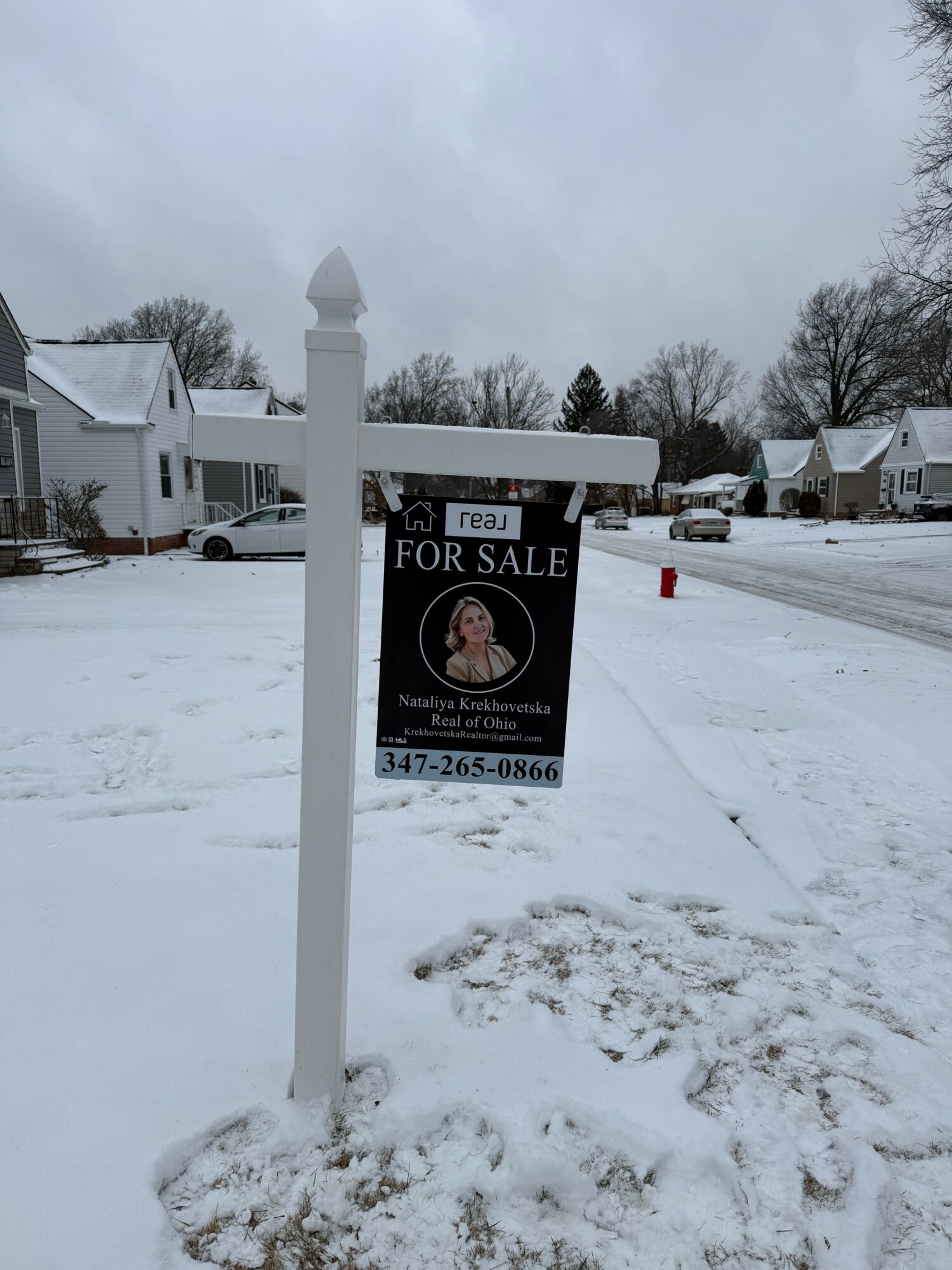 Snow-covered lot with a 'For Sale' sign, houses, and trees in the background, overcast sky.