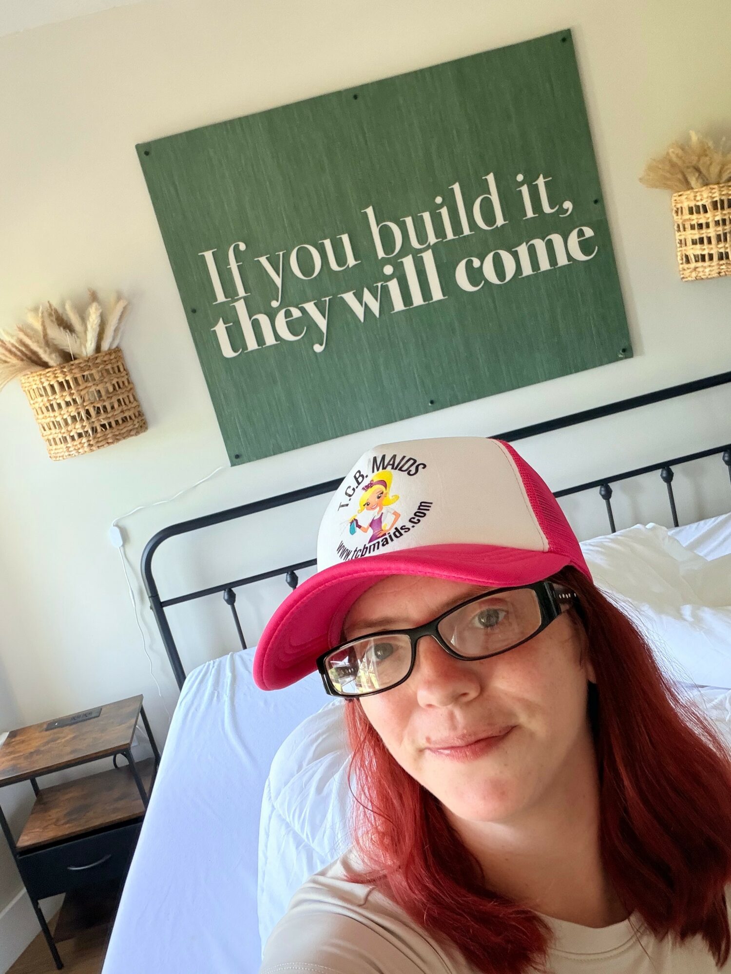 Person with red hair and glasses wearing a pink and white cap, smiling in a bedroom with a green sign on the wall.