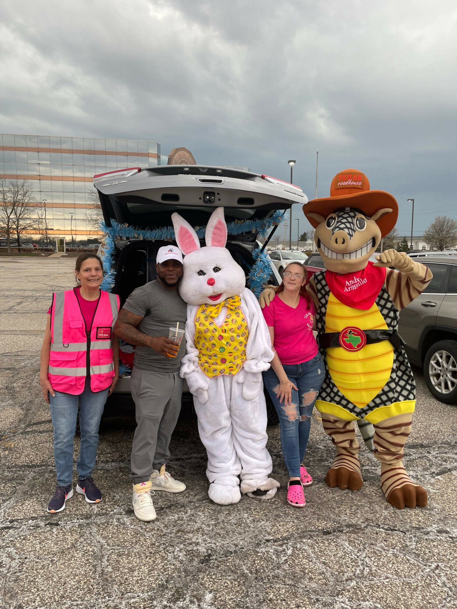 Group of people with a person in a bunny costume, standing outdoors near a car, with cloudy sky in background.