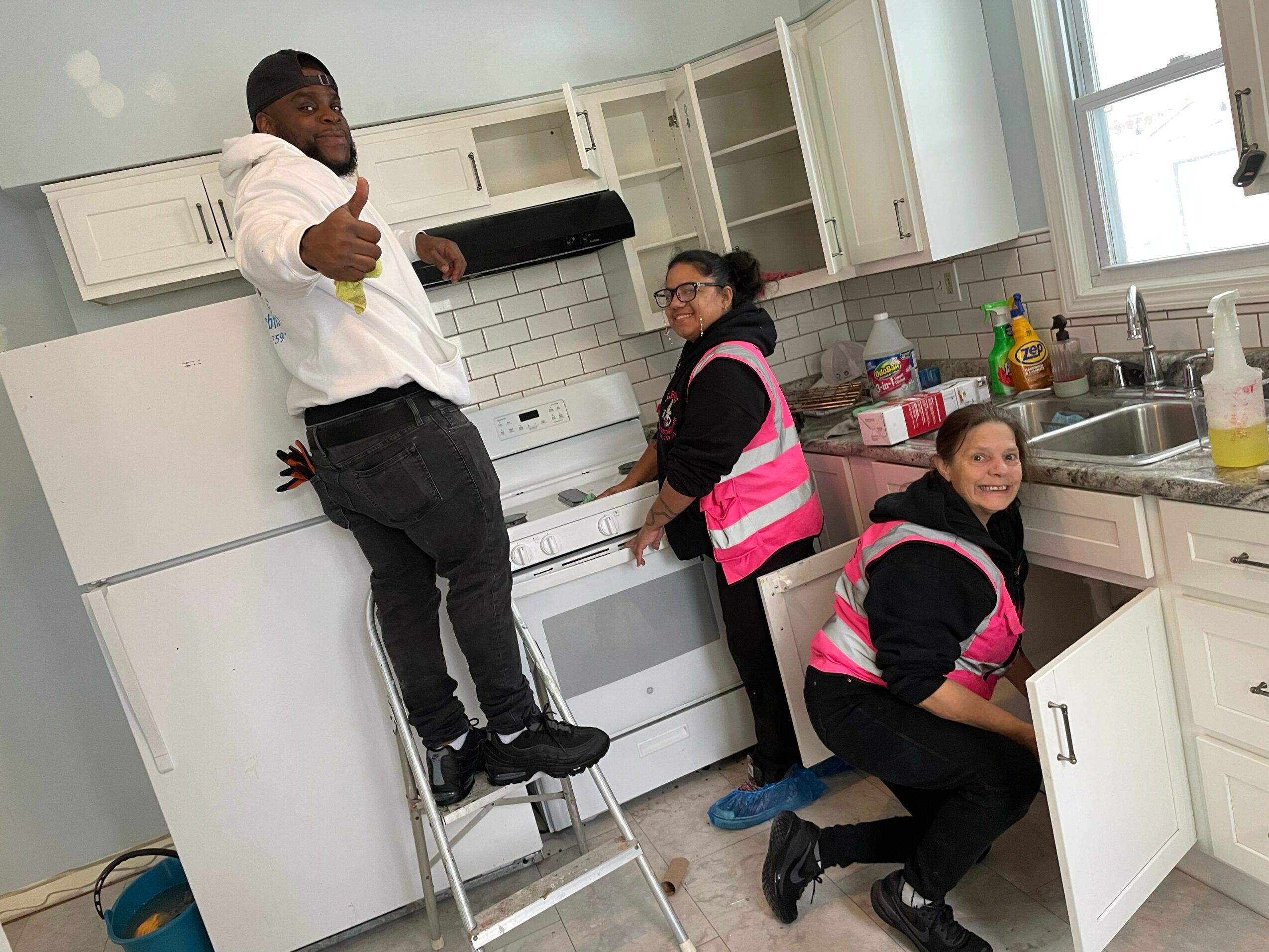 Three people in a kitchen, one standing on a stool giving a thumbs-up, two kneeling near the sink, smiling.