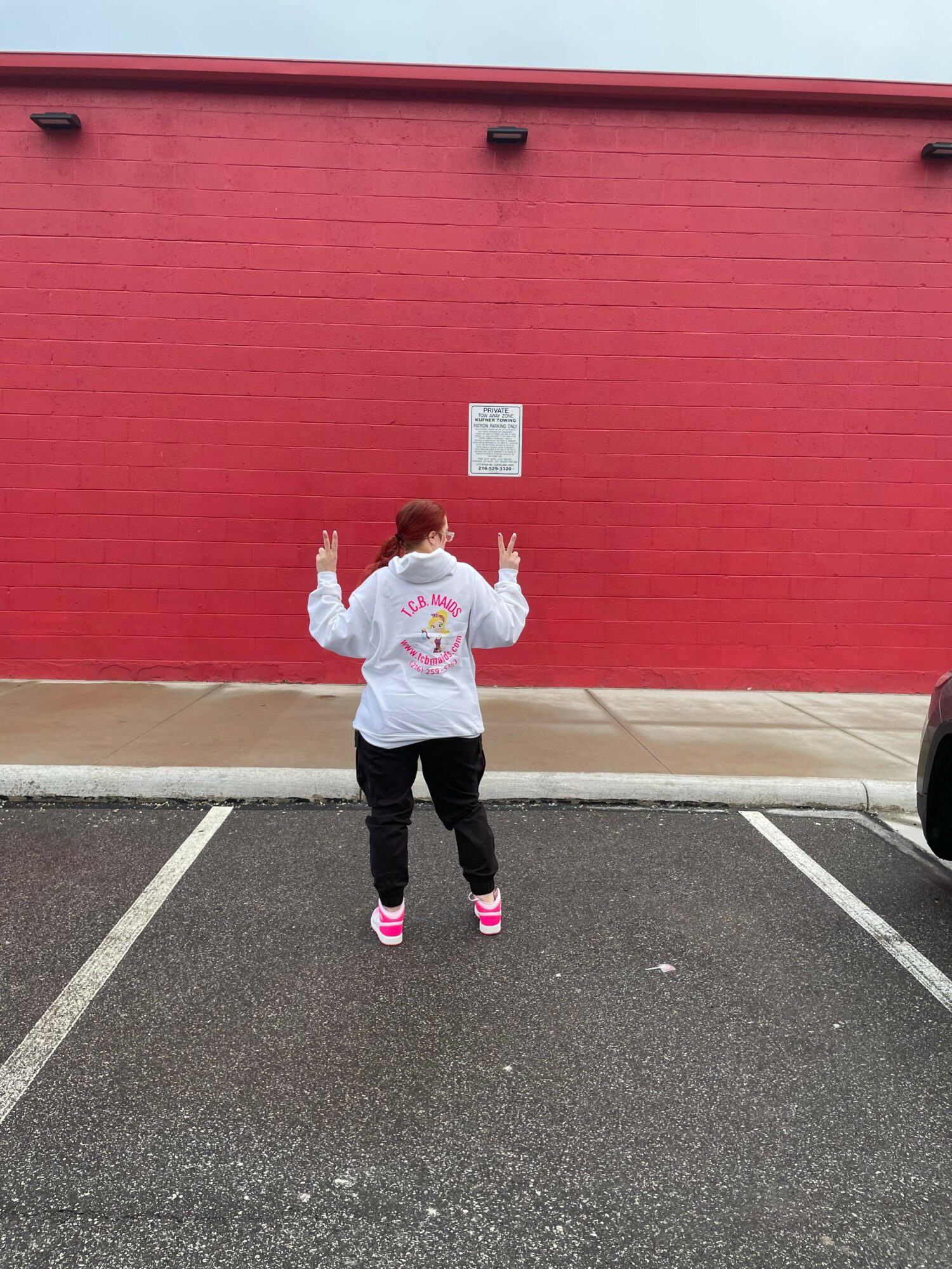 Person walking away from camera on sidewalk next to red wall, wearing white hoodie, black pants, and pink shoes.