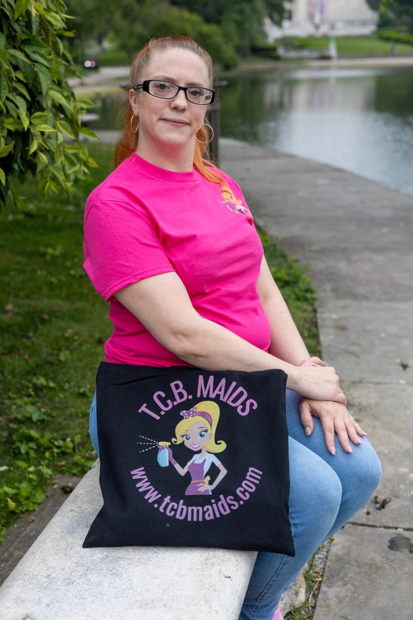 Woman with glasses and red hair sitting on a bench near water, holding a black tote bag with a logo.