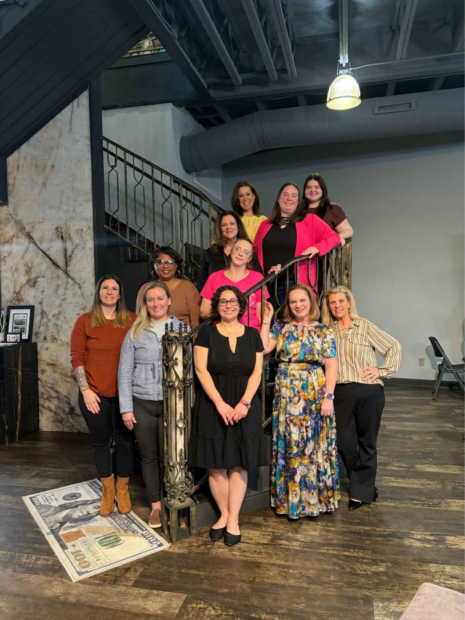 Group of ten women standing on a wooden floor in a room with a staircase, some smiling, dressed in casual and semi-formal clothing.