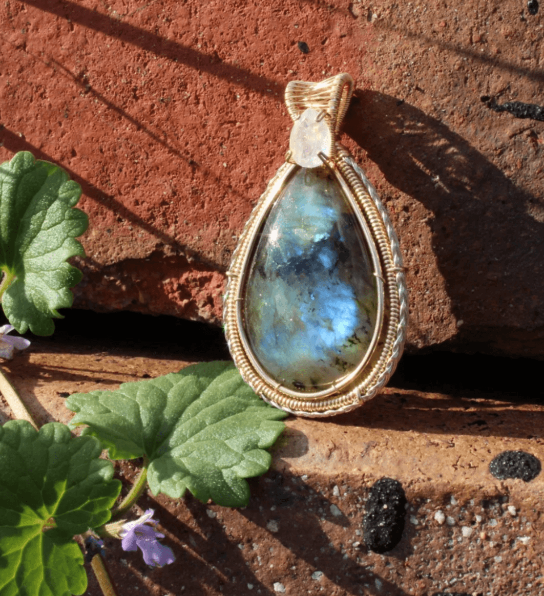 Pendant with a teardrop-shaped blue gemstone, gold wire wrapping, resting on a brick surface with green leaves nearby.