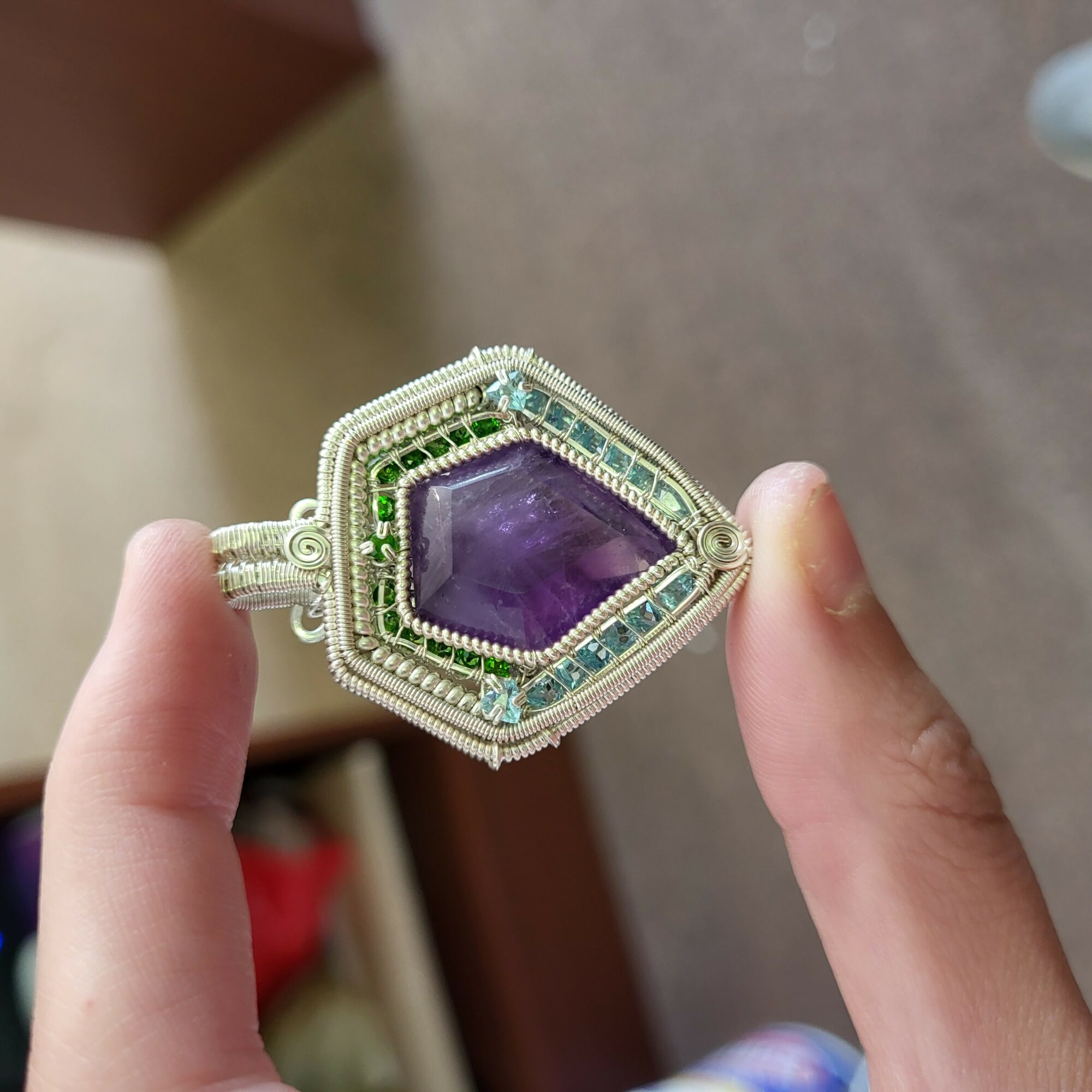 Person holding a silver ring with a large purple gemstone and green accents, against a blurred background.