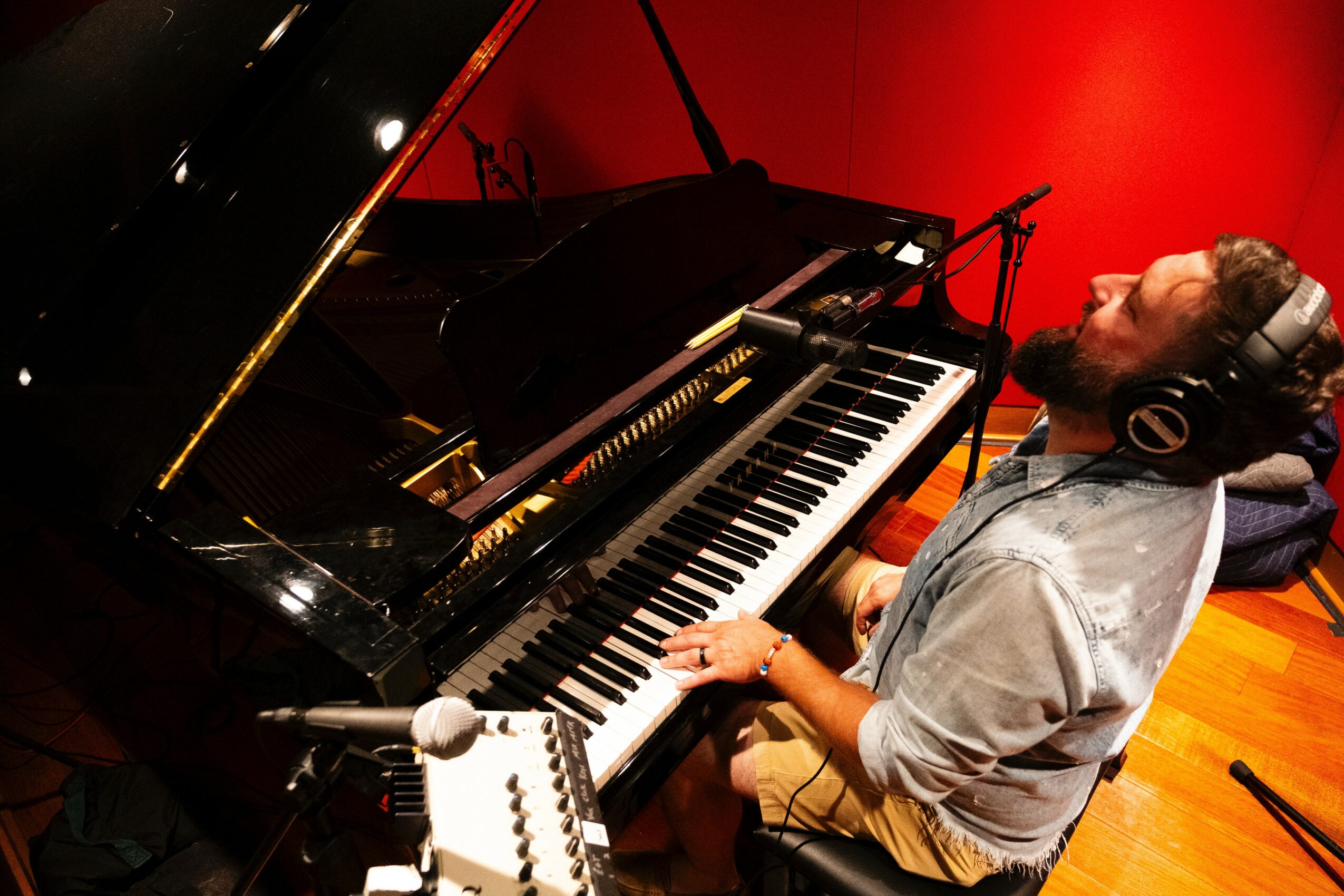 Person playing grand piano in recording studio with red wall, wearing headphones, focused on music, wooden floor, microphone nearby.