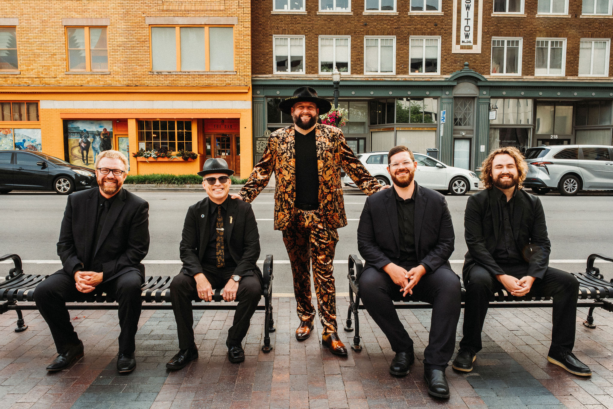 Five men sitting and standing on a city sidewalk in front of buildings and parked cars, smiling, dressed in dark and patterned clothing.