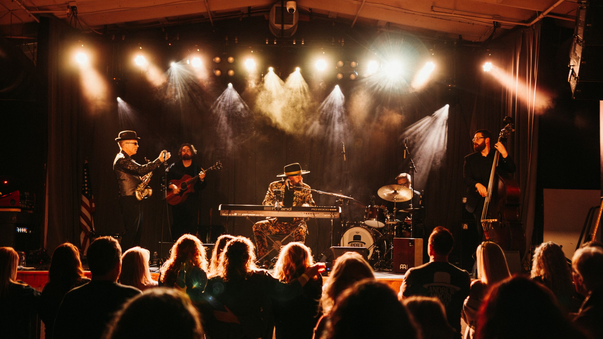 Musicians perform on stage with bright lights, audience watching in a darkened venue, some silhouettes visible.
