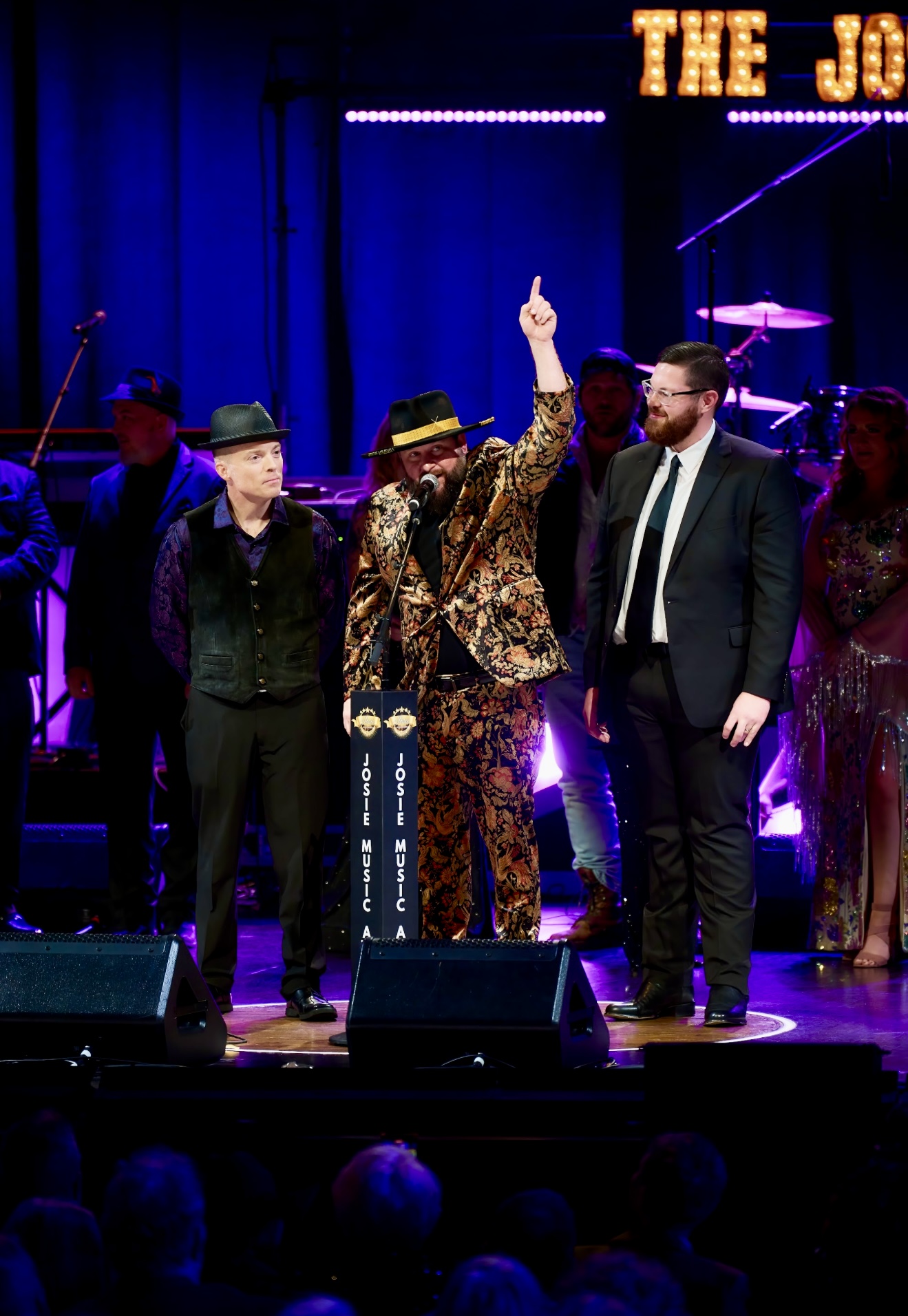 Three men on stage, one in a leopard print suit raising a finger, others in dark suits, with a band and audience in background.