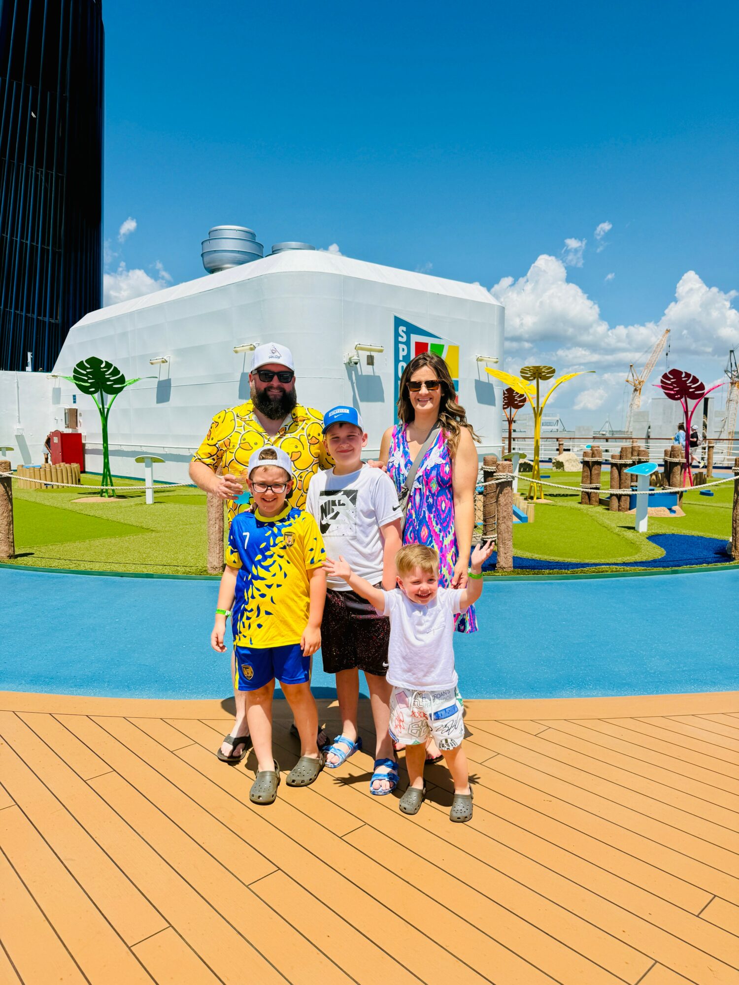 Family of five standing on a wooden deck with a colorful amusement park in the background, including a large white structure and playful trees.