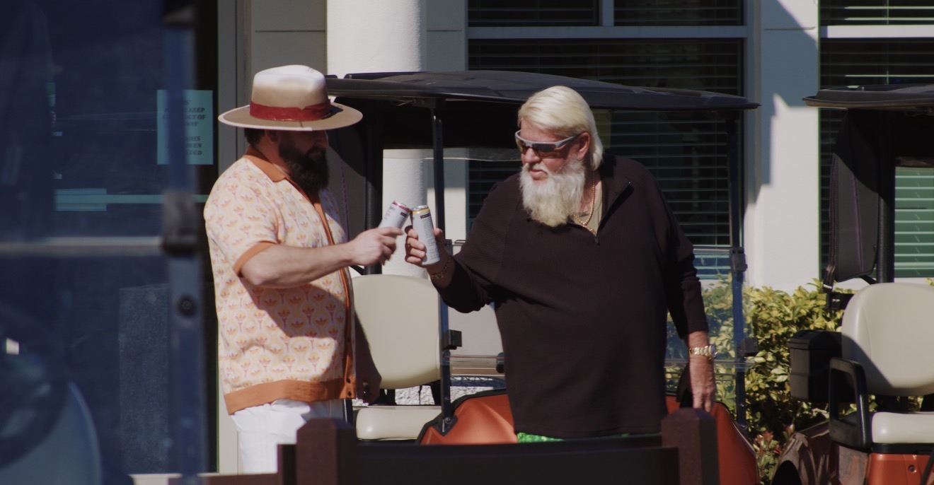 Two people exchanging a drink outdoors, one wearing a hat and the other with a beard, in front of a building.