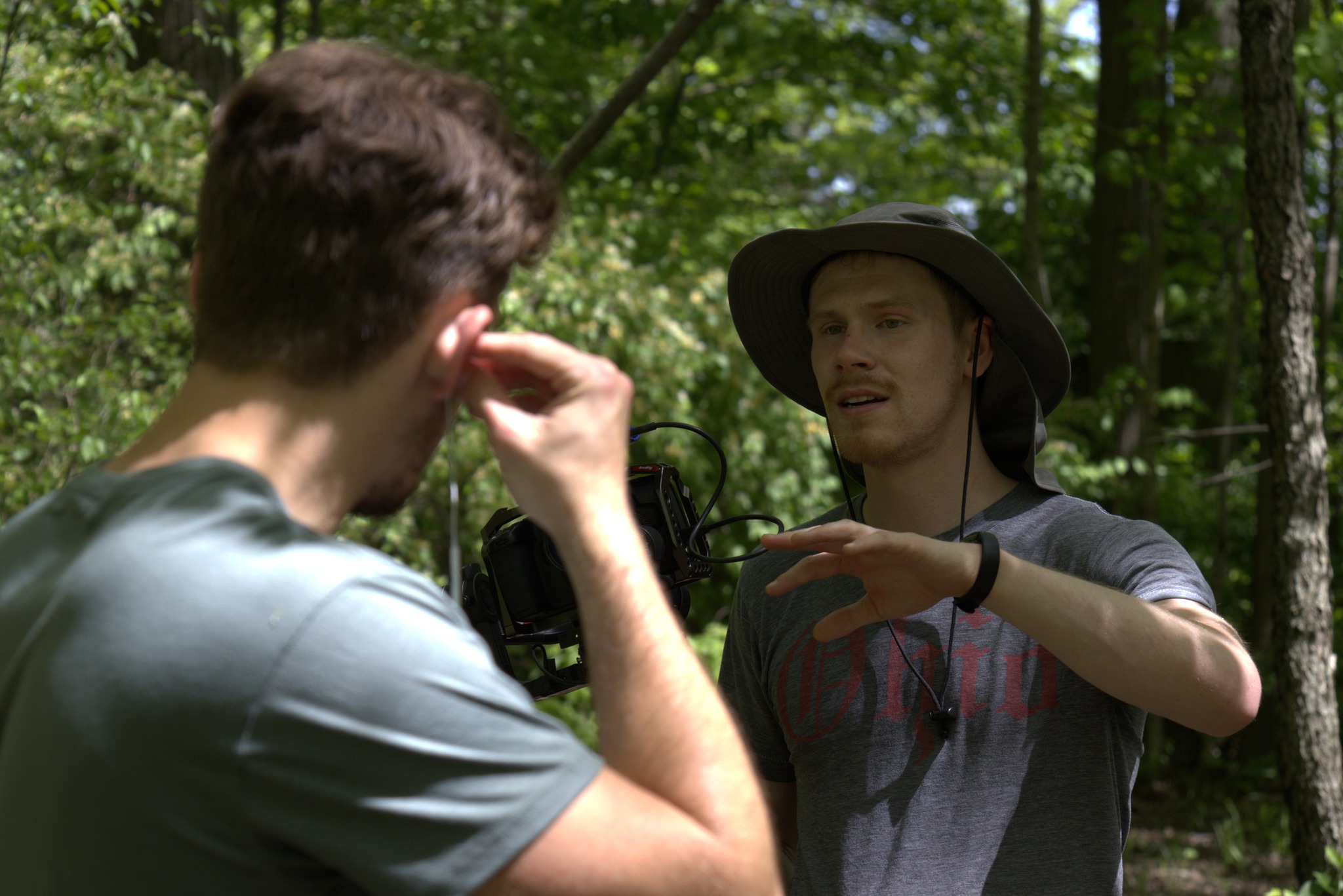 Two people talking outdoors in a wooded area, one wearing a hat and gesturing with hands, the other listening.