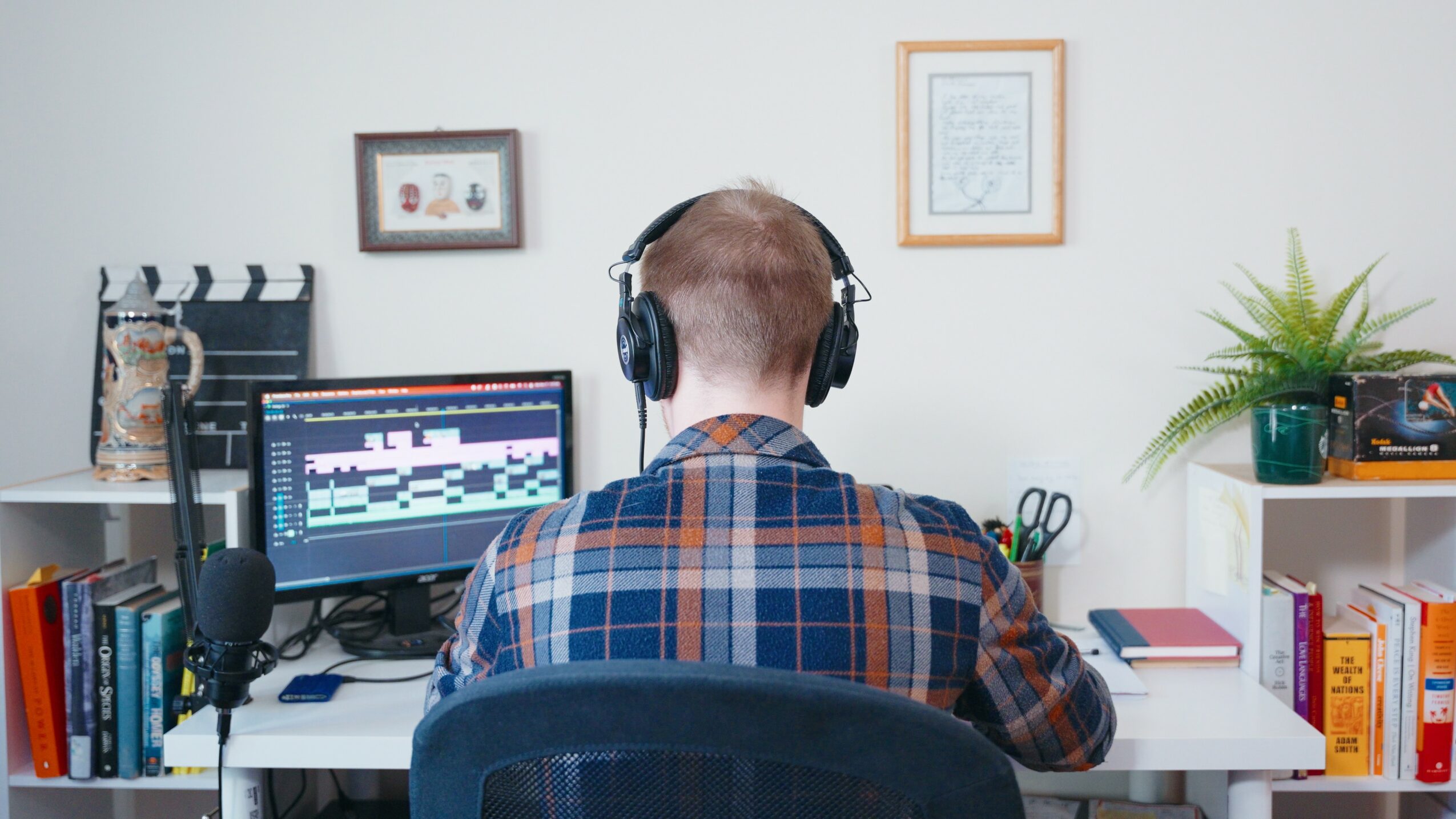 Person with headphones working on a computer in a home office, with books, plants, and framed pictures on the wall.
