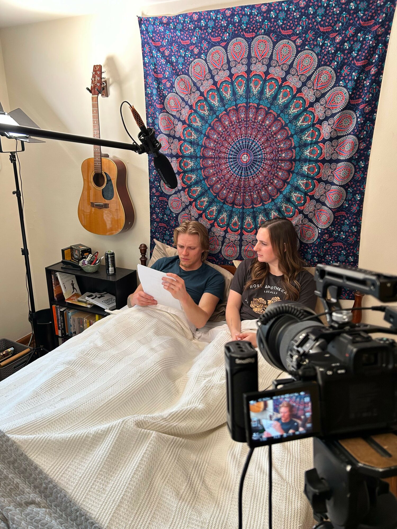 Person filming a woman sitting on a bed with a colorful mandala tapestry behind her, guitar on the wall, and camera equipment present.