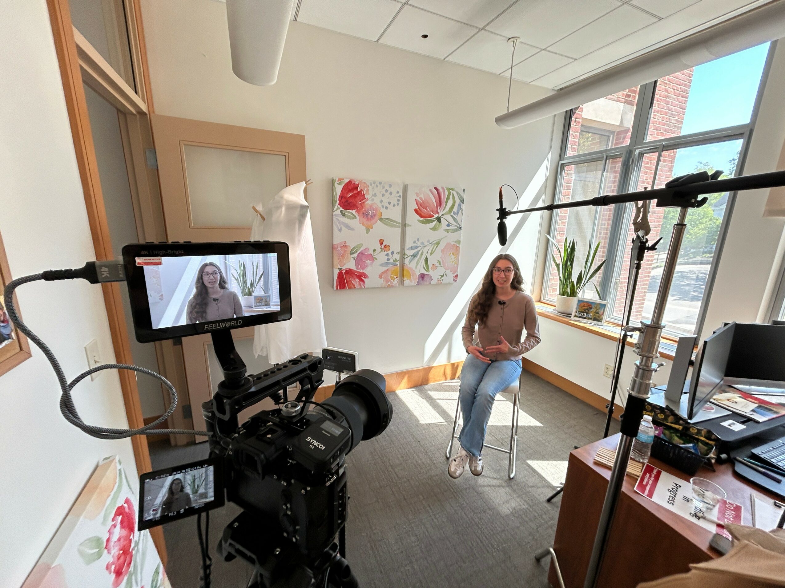 Woman sitting on a chair in a room with large window, plants, and artwork, being filmed with professional camera equipment.