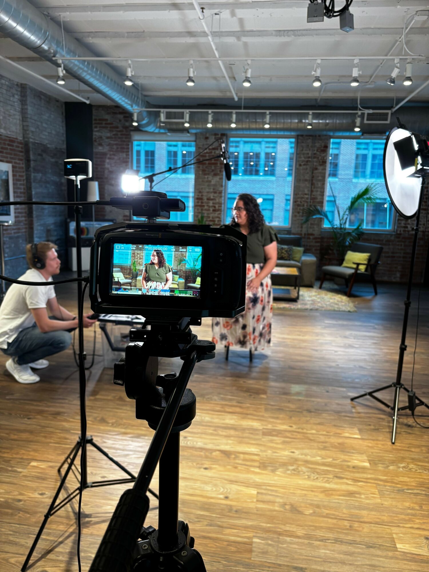 Person standing in a room with wooden floor, large windows, and a camera on a tripod capturing the scene.
