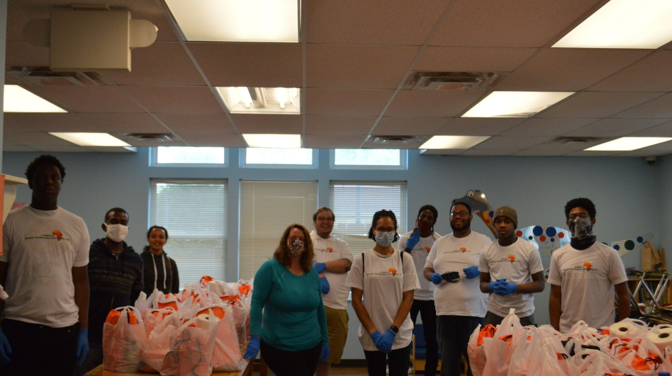 Group of people standing in a room with windows, some wearing masks, surrounded by bags or packages.