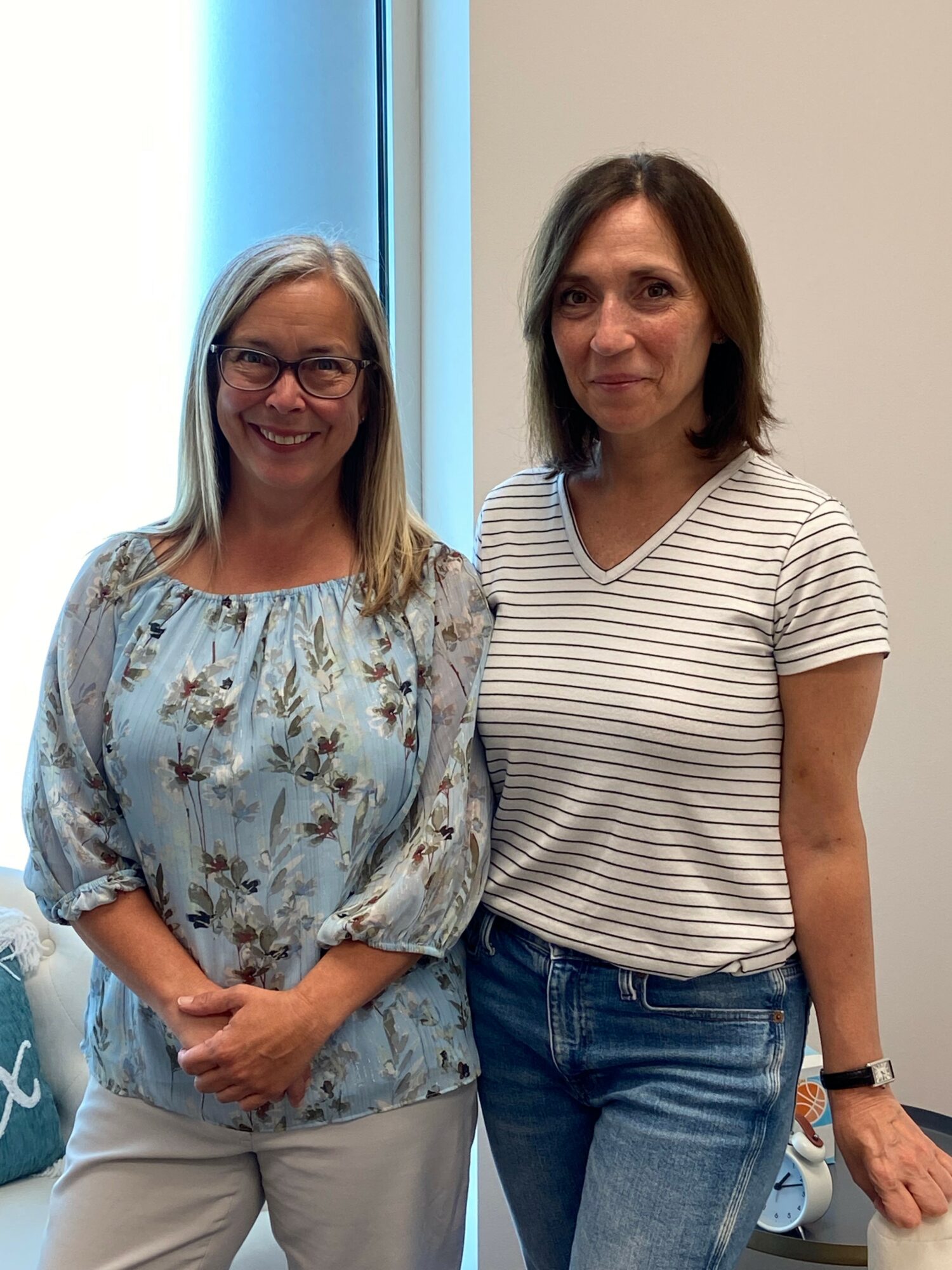 Two women standing indoors, one smiling and wearing glasses, the other with a neutral expression, both facing forward.