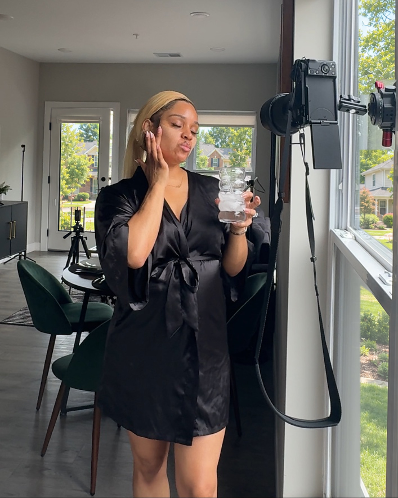 Woman in black robe holding a water bottle, looking at a camera mounted on a window, in a bright room with dining area.