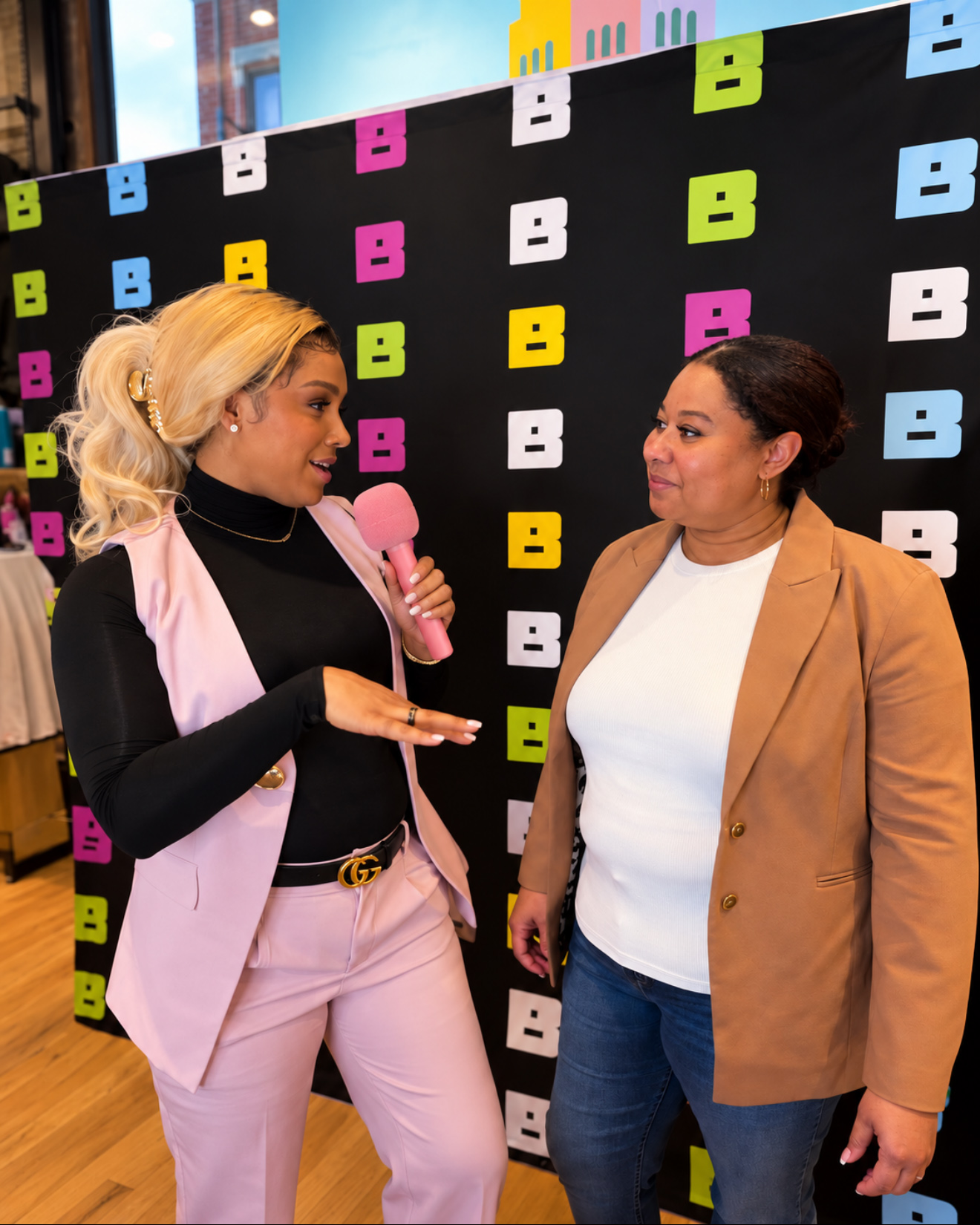 Two women talking in front of a colorful backdrop with letters B in pink, blue, green, and yellow, at an indoor event.