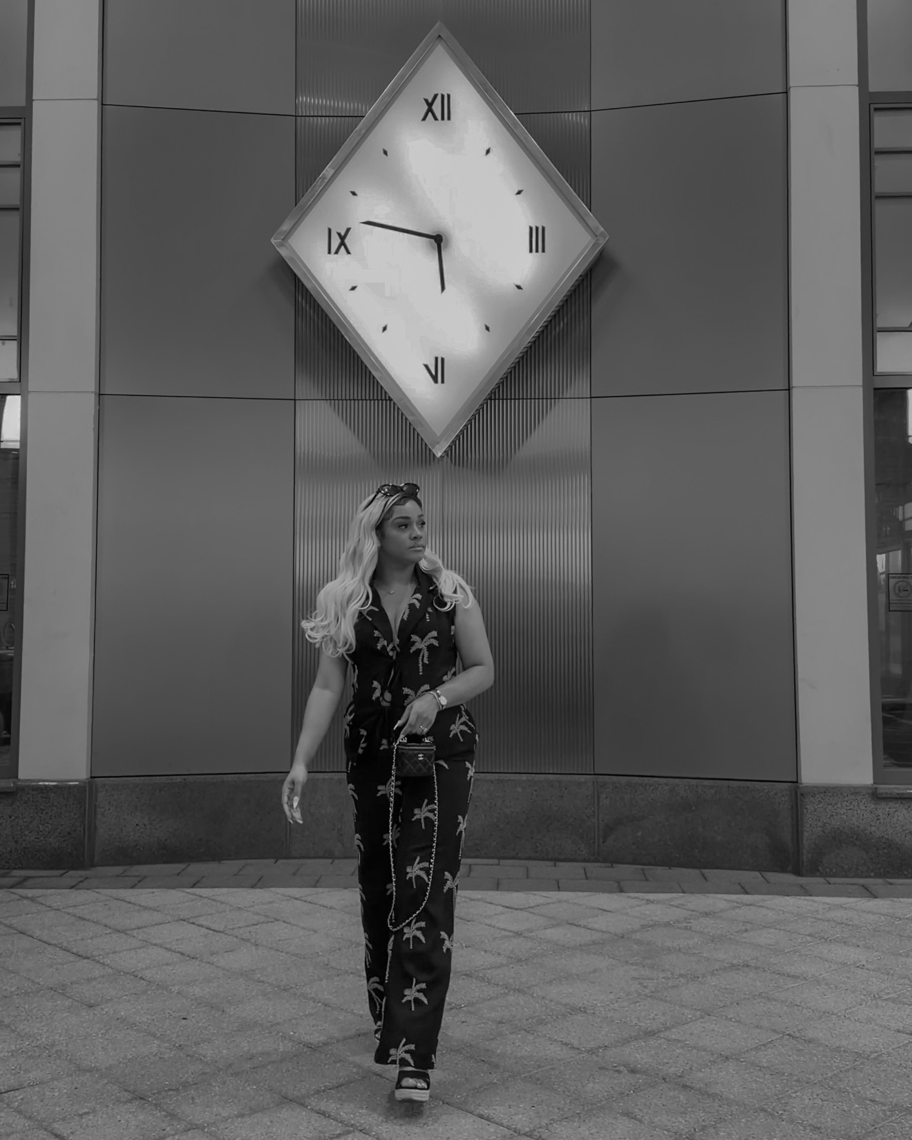 Woman walking in front of a wall with a large, tilted clock showing 9:45, on a city sidewalk.