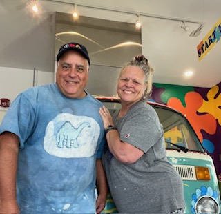 Two smiling people standing indoors, one wearing a blue shirt and the other in a gray shirt, with colorful wall art behind them.