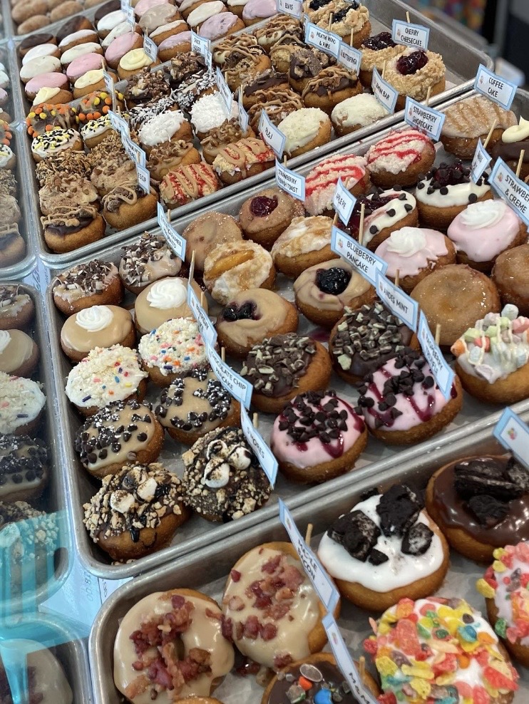 Display of various decorated donuts and pastries with small labels, arranged in rows on trays.