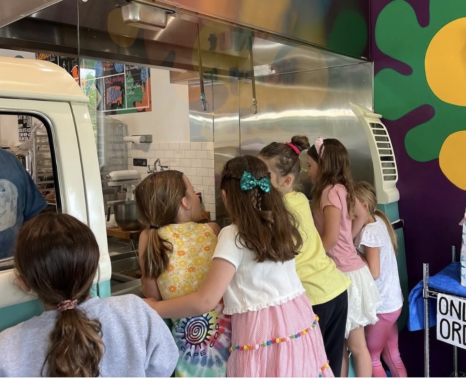 Group of children waiting in line at a colorful food truck, some talking and looking inside.