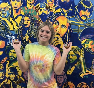 Young woman smiling with arms raised in front of a colorful mural of diverse faces.