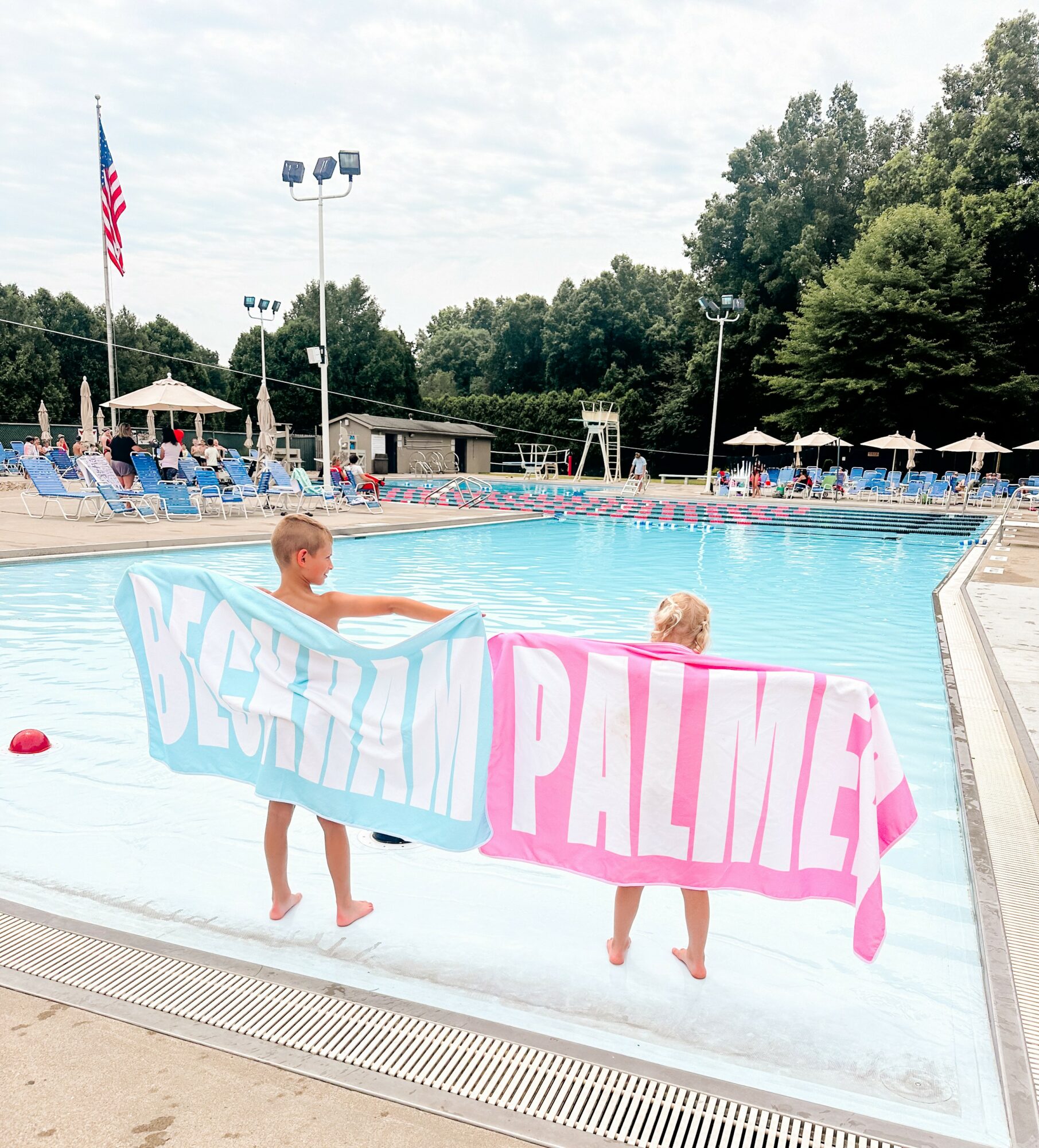 Two children hold a large towel with the words 'YOUTH' and 'PALM' at a swimming pool, with lounge chairs and trees in background.