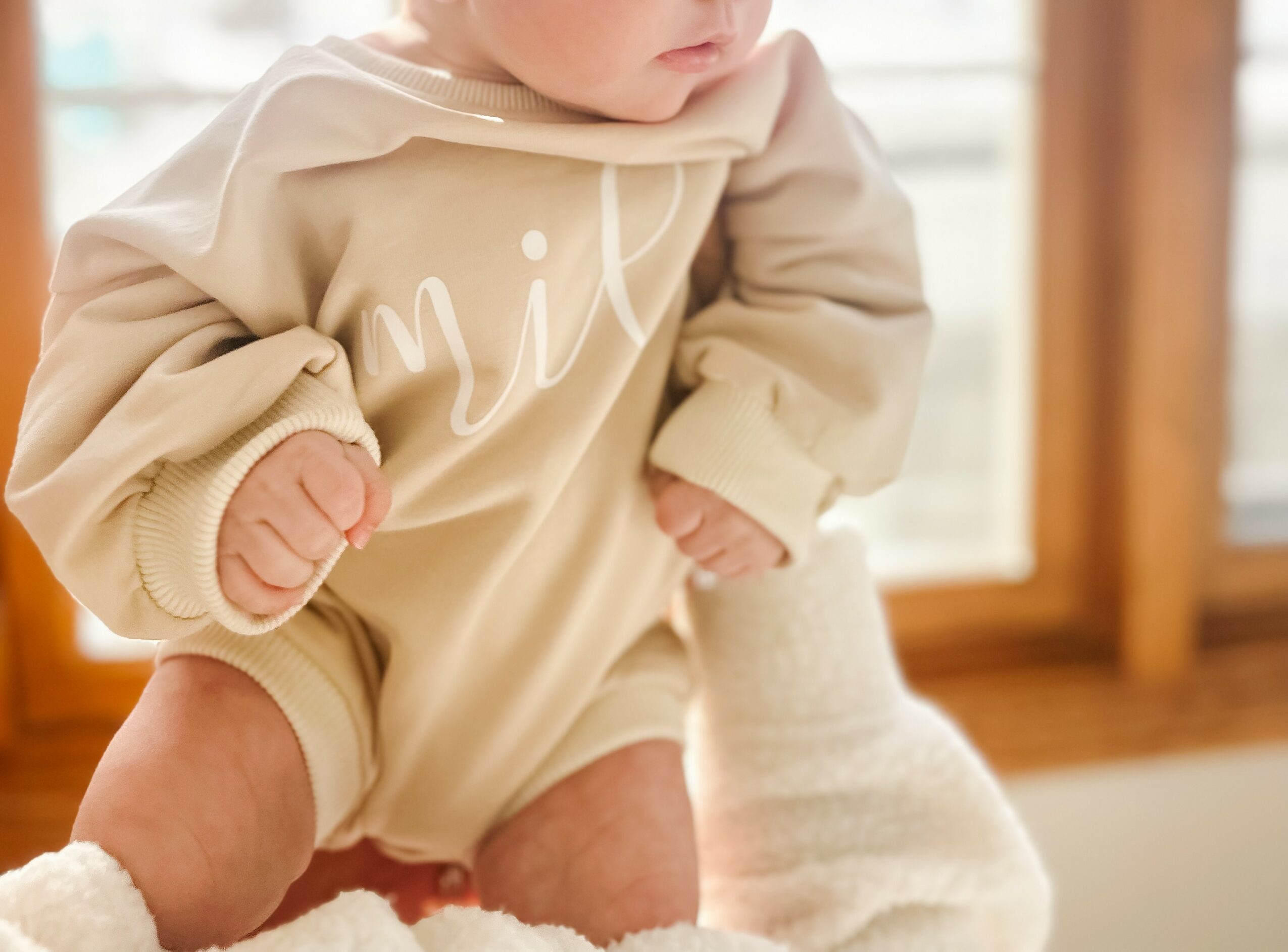 Close-up of a baby in a beige hoodie, sitting on a soft surface near a window, with wooden furniture in background.