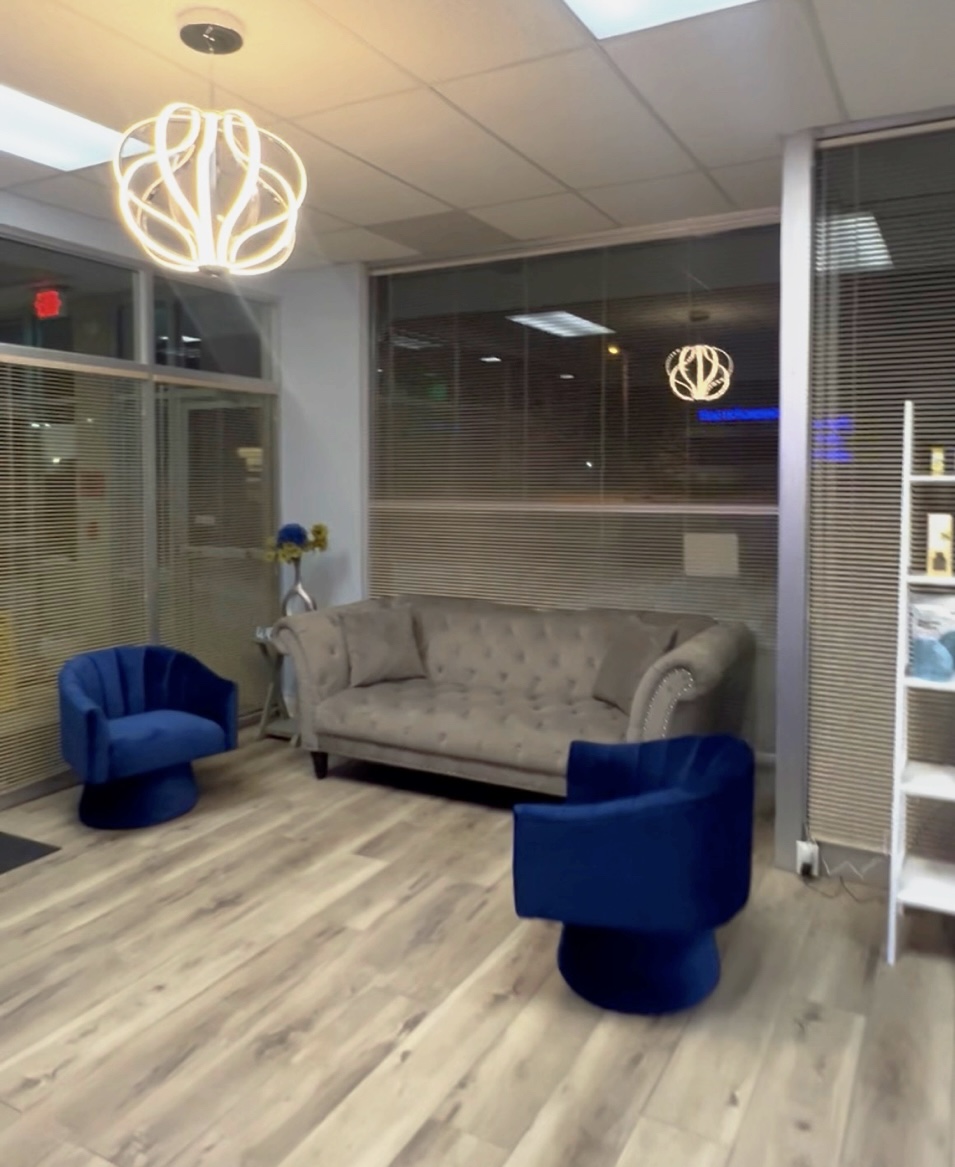 Interior space with a gray sofa, two blue chairs, a glass wall, and modern lighting fixtures, wood flooring, and a window.