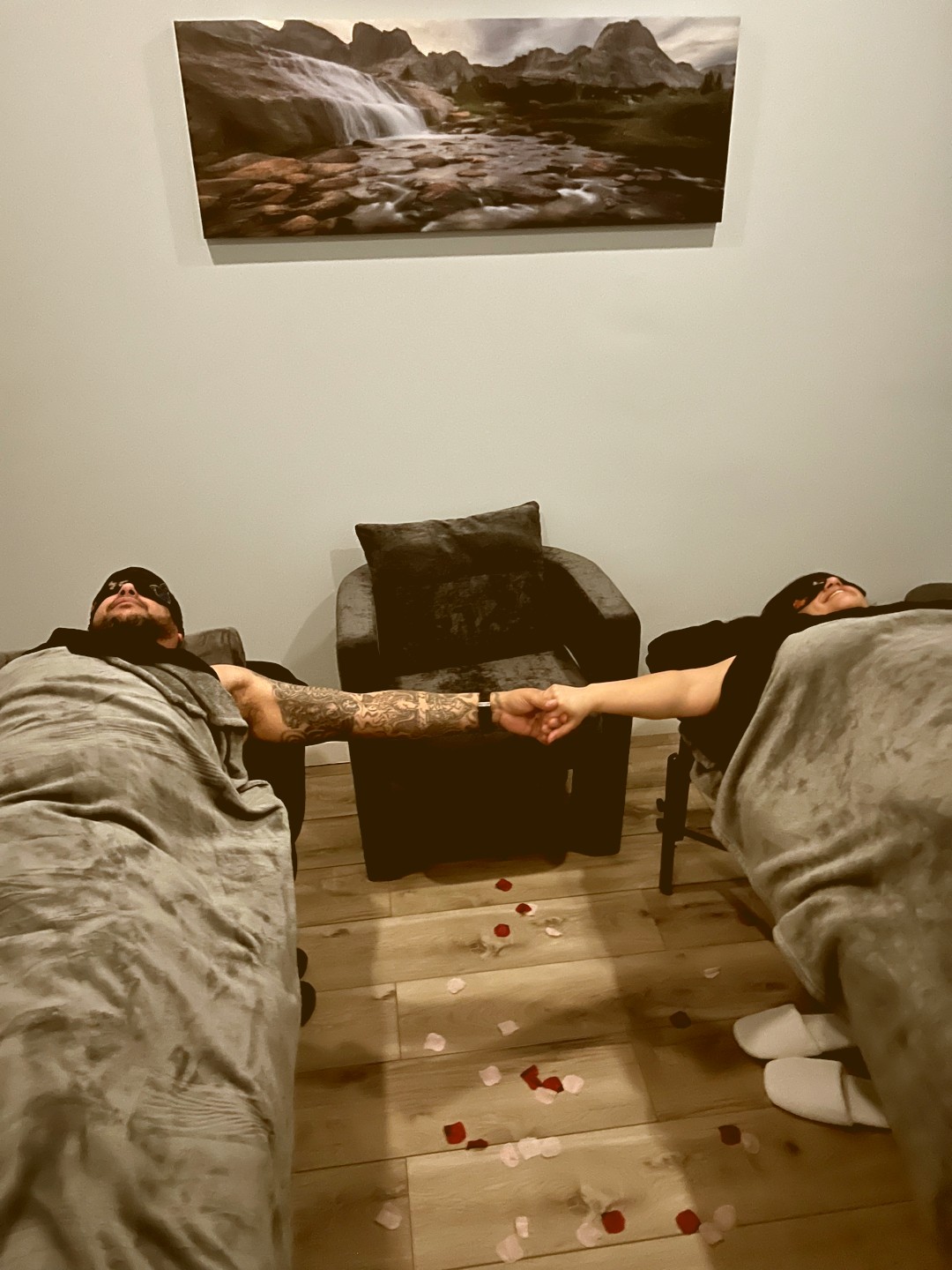 Two people lying on beds holding hands, with a painting of a waterfall on the wall behind them, and bloodstains on the floor.