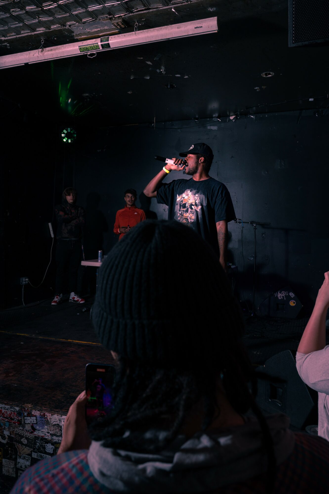 Person holding a microphone on stage with a dark background, audience in foreground, and stage lighting.