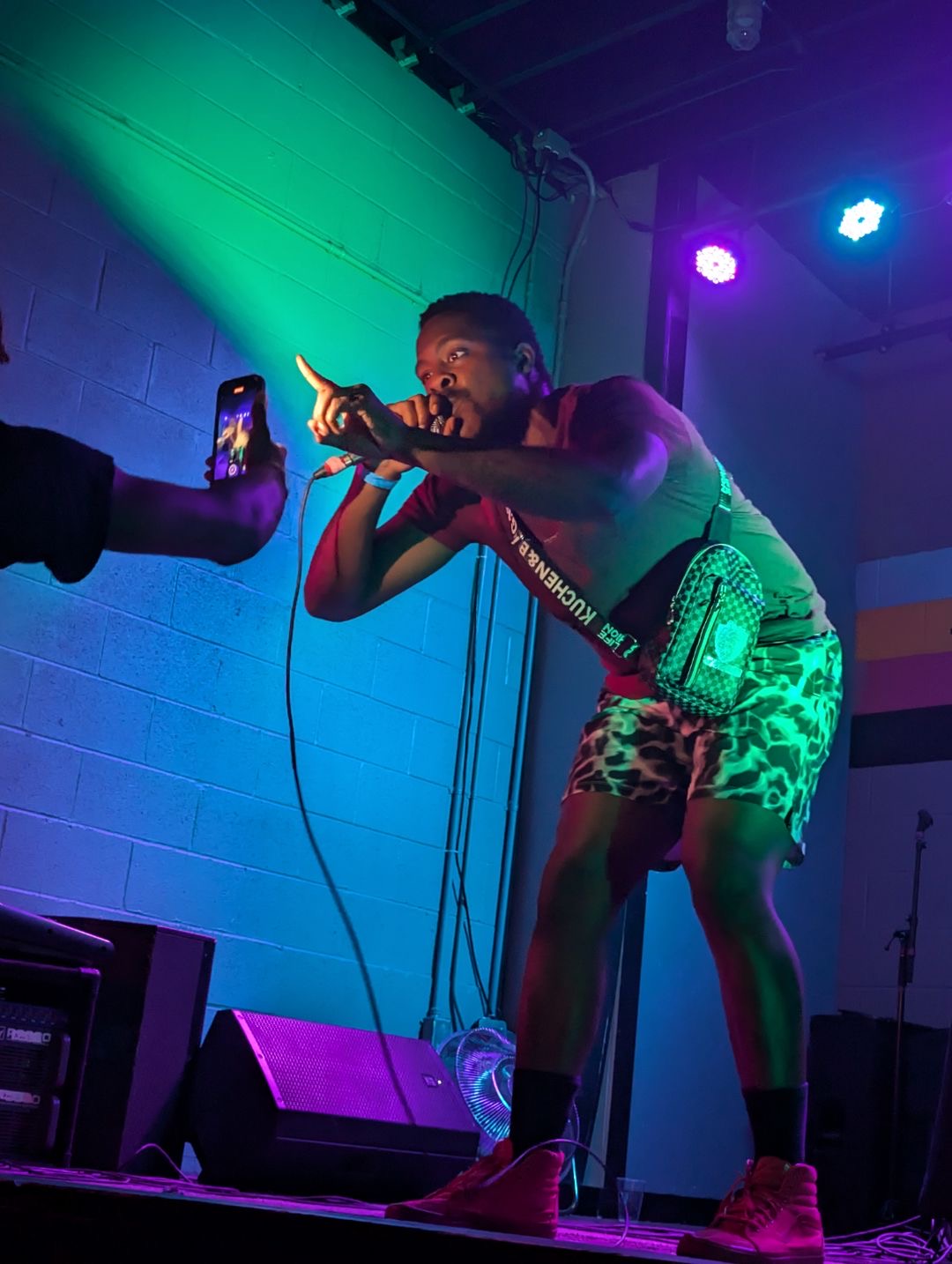 Performer singing into microphone on stage with colorful lighting, holding a phone, wearing patterned shorts and red shoes.