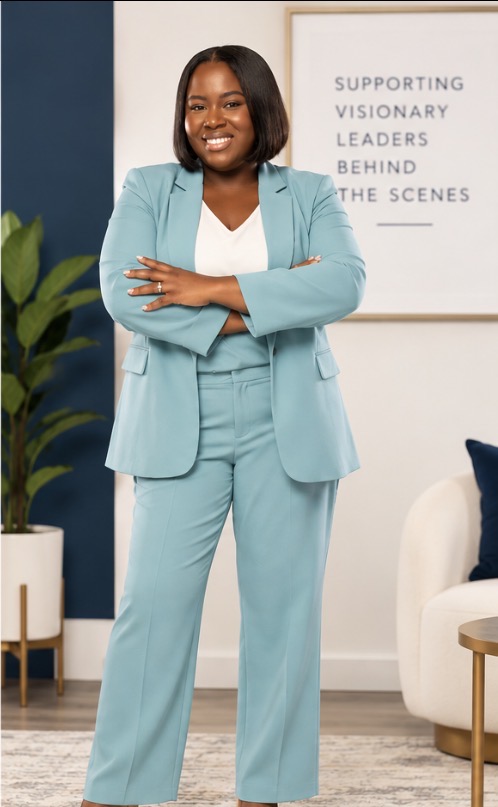 Woman in a light blue suit standing with arms crossed in a modern office space, smiling, with a framed quote on the wall.