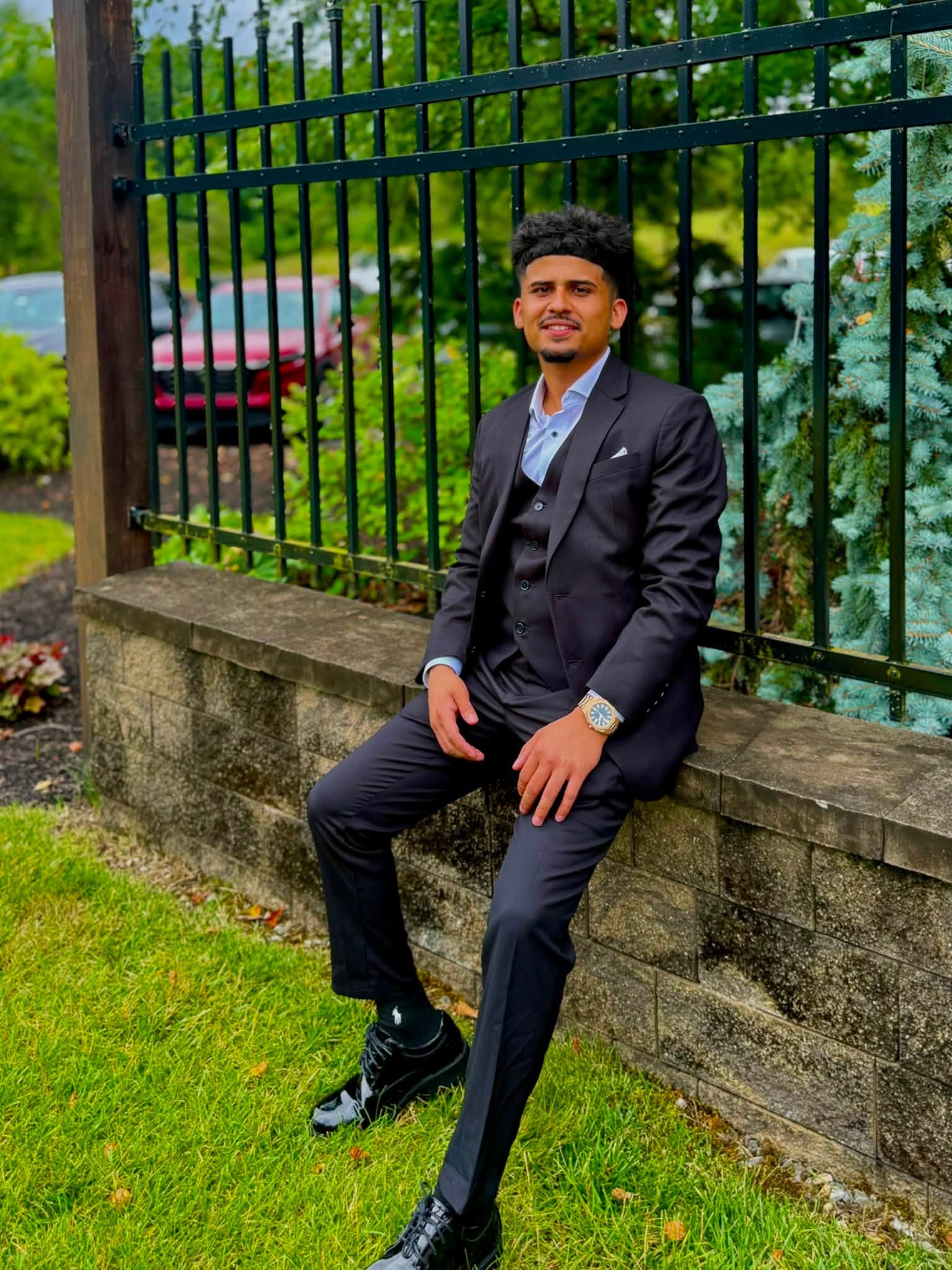Man in a suit sitting on a stone ledge next to a black metal fence in a park, greenery in background.
