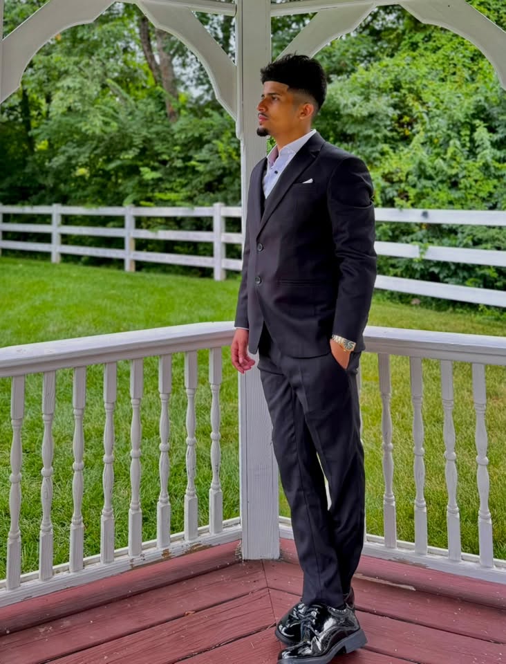 Young man in a dark suit standing on a white gazebo with green trees and grass in background.