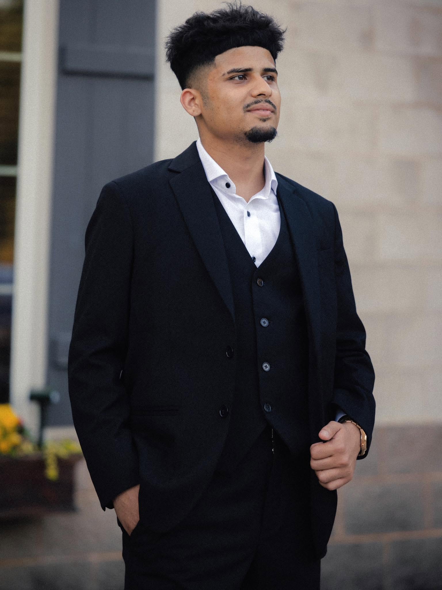 Young man in formal suit standing outdoors, looking to the side, with a neutral background.
