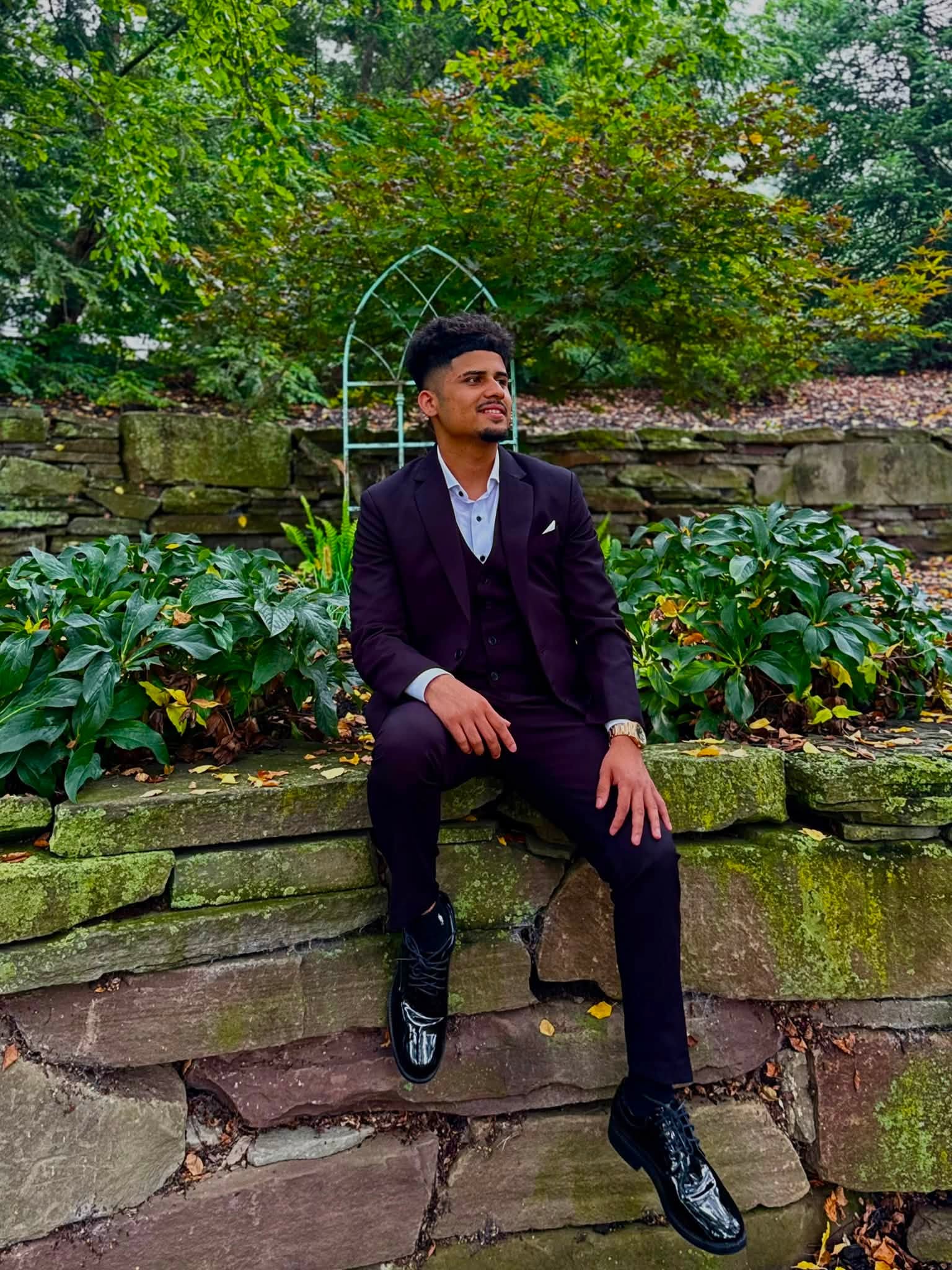 Young man in formal suit sitting on stone steps outdoors, surrounded by greenery and plants, with a decorative arch behind him.